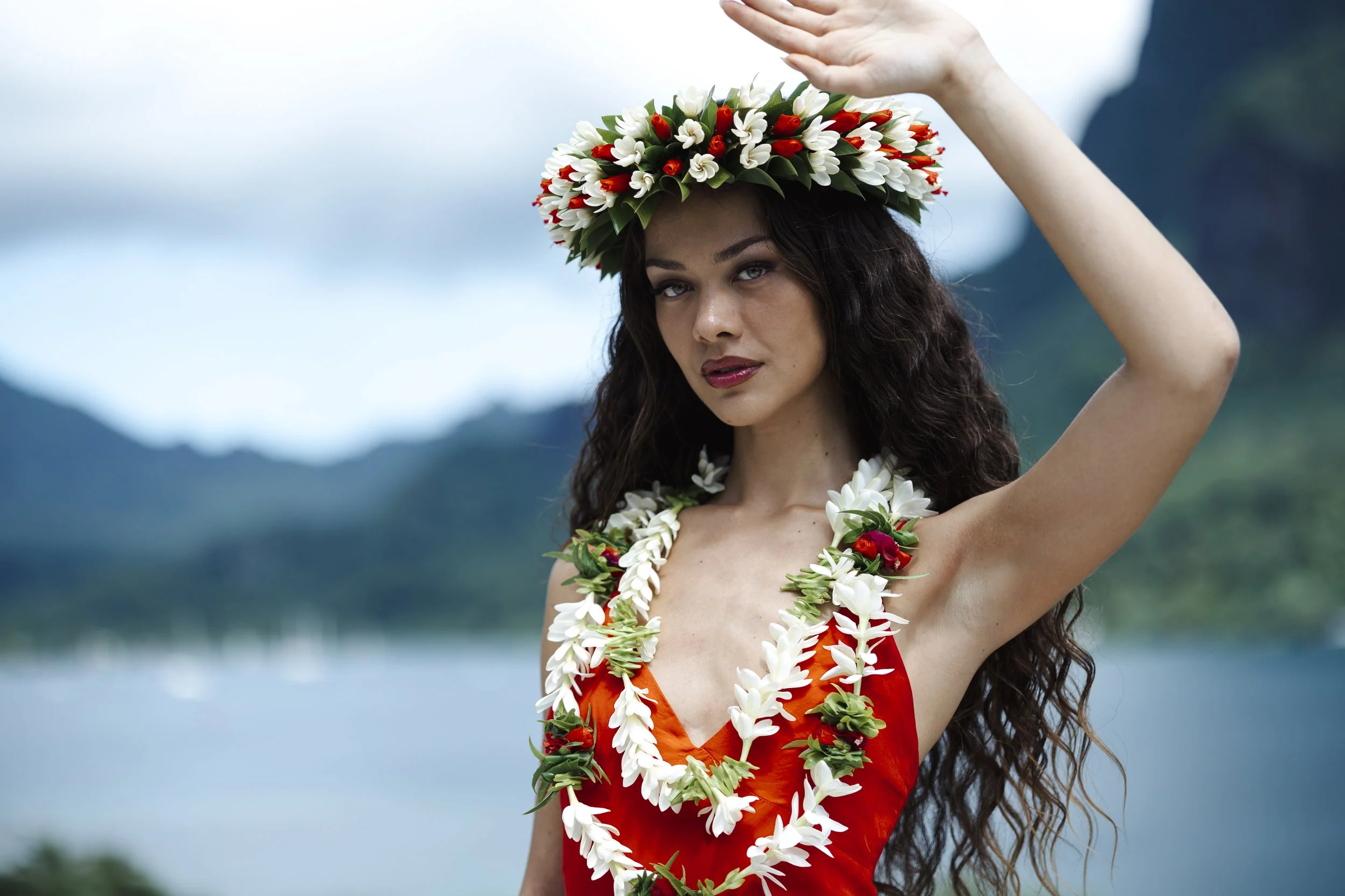 Jeune femme portant une couronne de fleurs blanches et rouges, un collier de fleurs blanches et rouges, et une robe rouge, devant un lac et des montagnes.