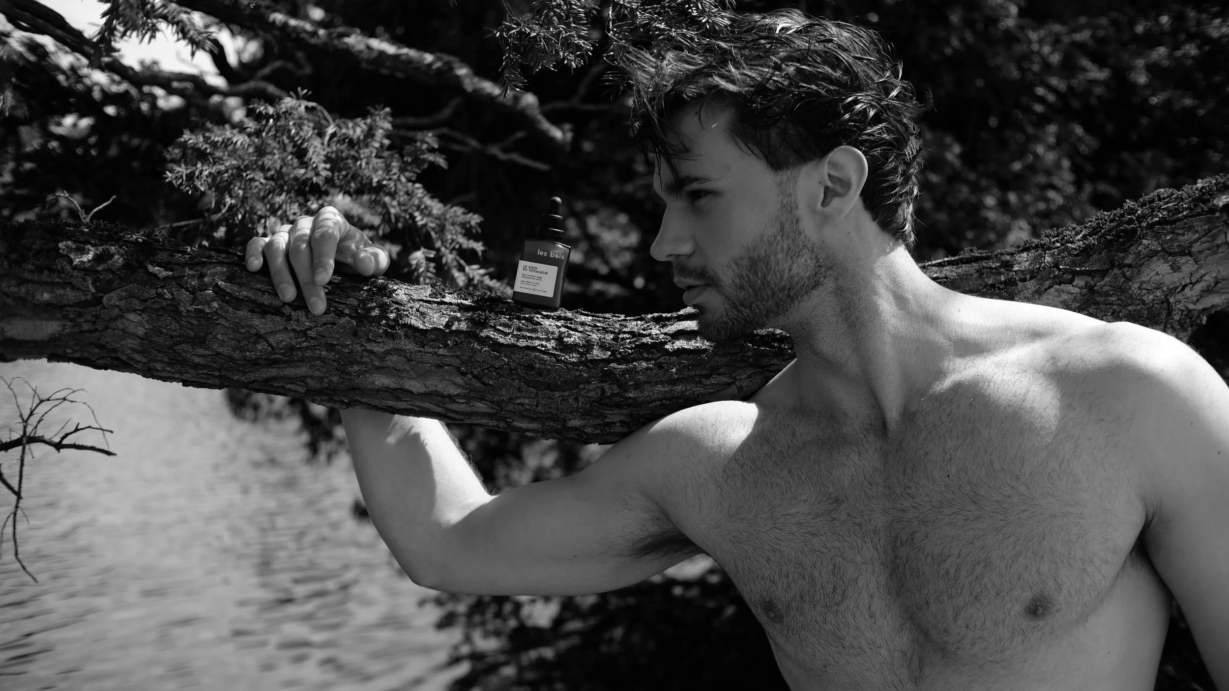 Un homme nu portant un arbre sur l'épaule, dans un cadre naturel avec des arbres et de l'eau en arrière-plan, en noir et blanc.