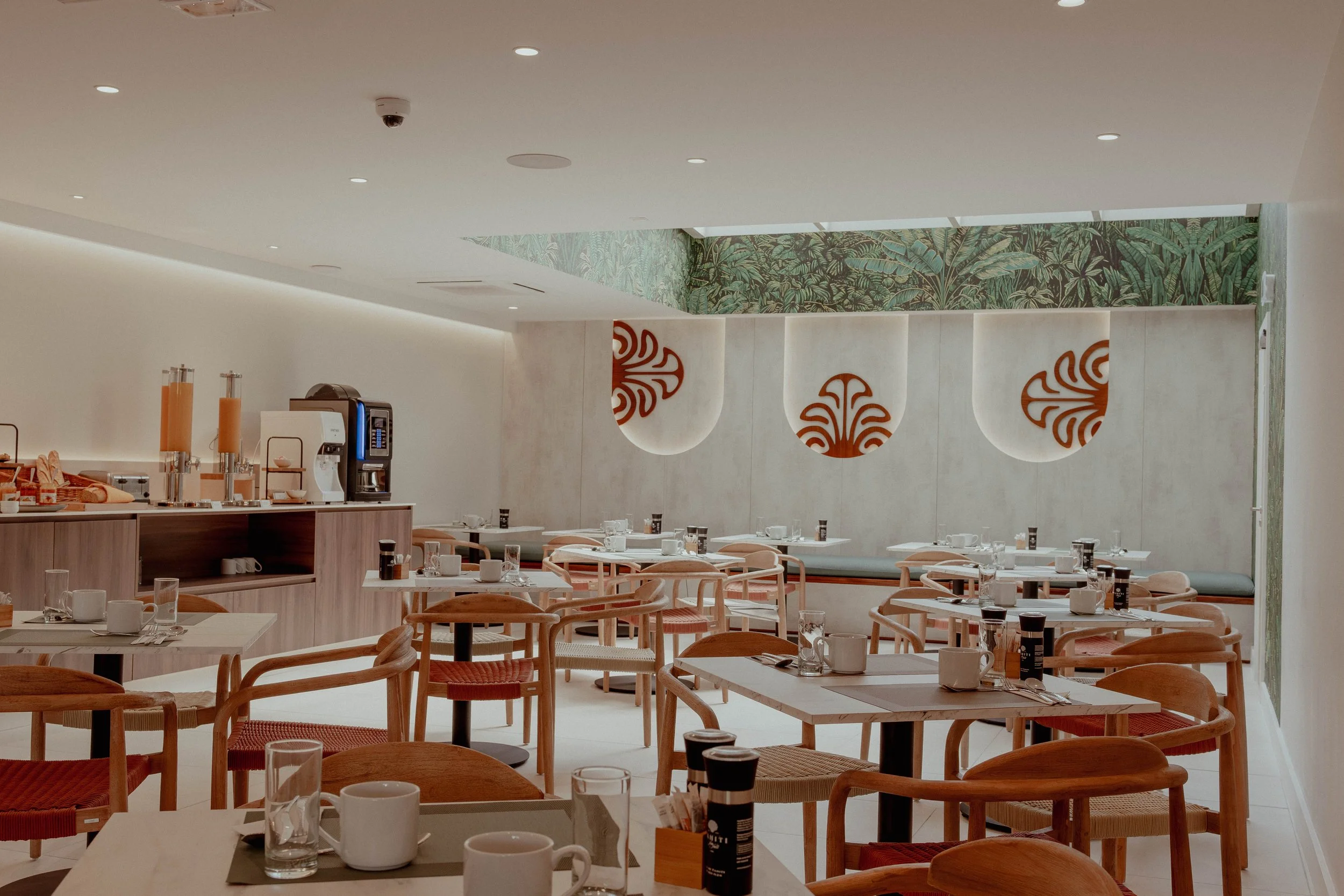 Salle à manger d'un restaurant avec des tables en bois, des chaises en bois avec assises en tissu rouge, des tasses, des verres, des assiettes, et un buffet avec des distributeurs de jus et une machine à café. Mur blanc avec des décorations en forme 