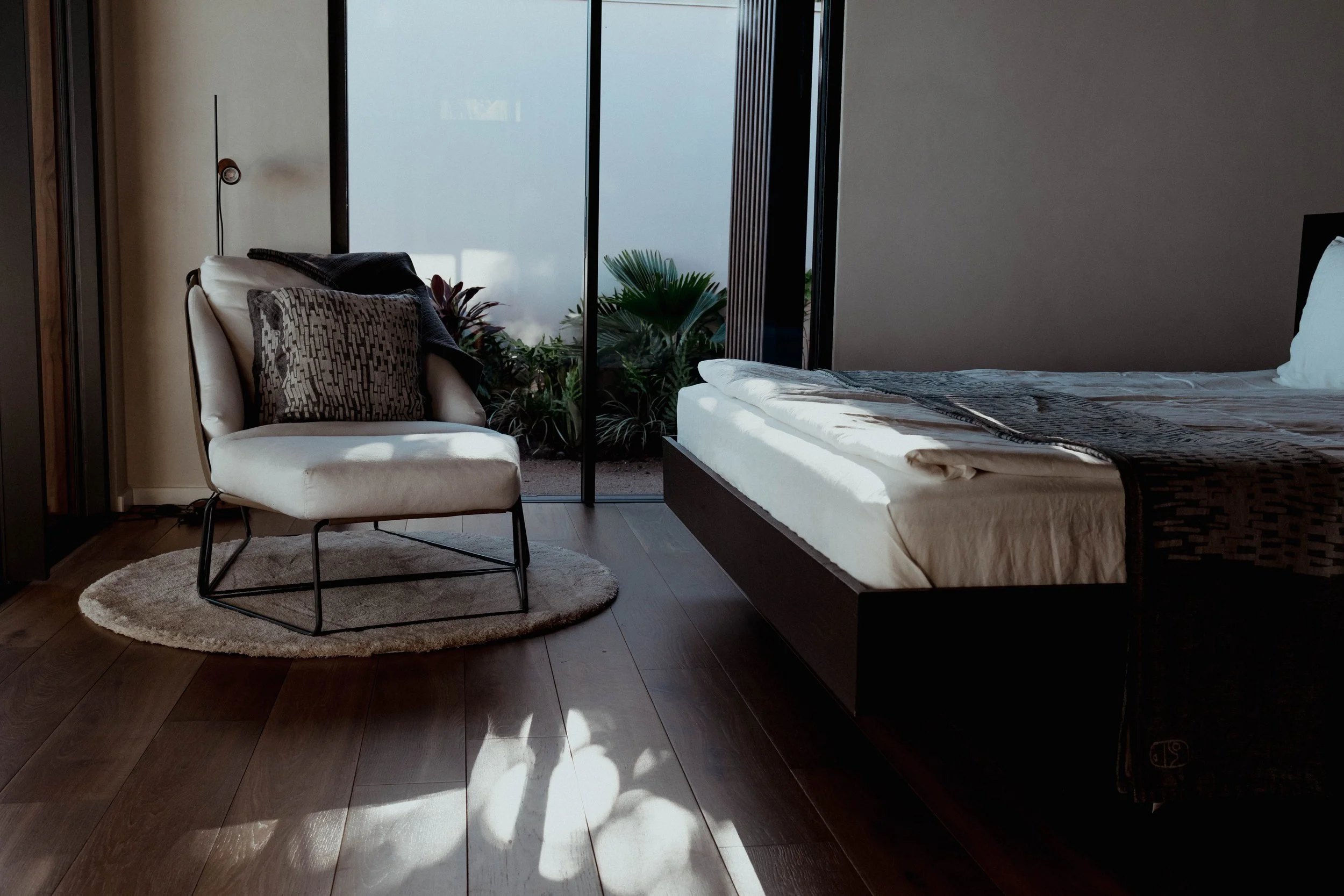 Chambre avec un lit, une chaise beige avec coussin, une lampe de lecture, un tapis rond, une grande fenêtre en verre laissant entrer la lumière naturelle, des plantes vertes à l'extérieur.