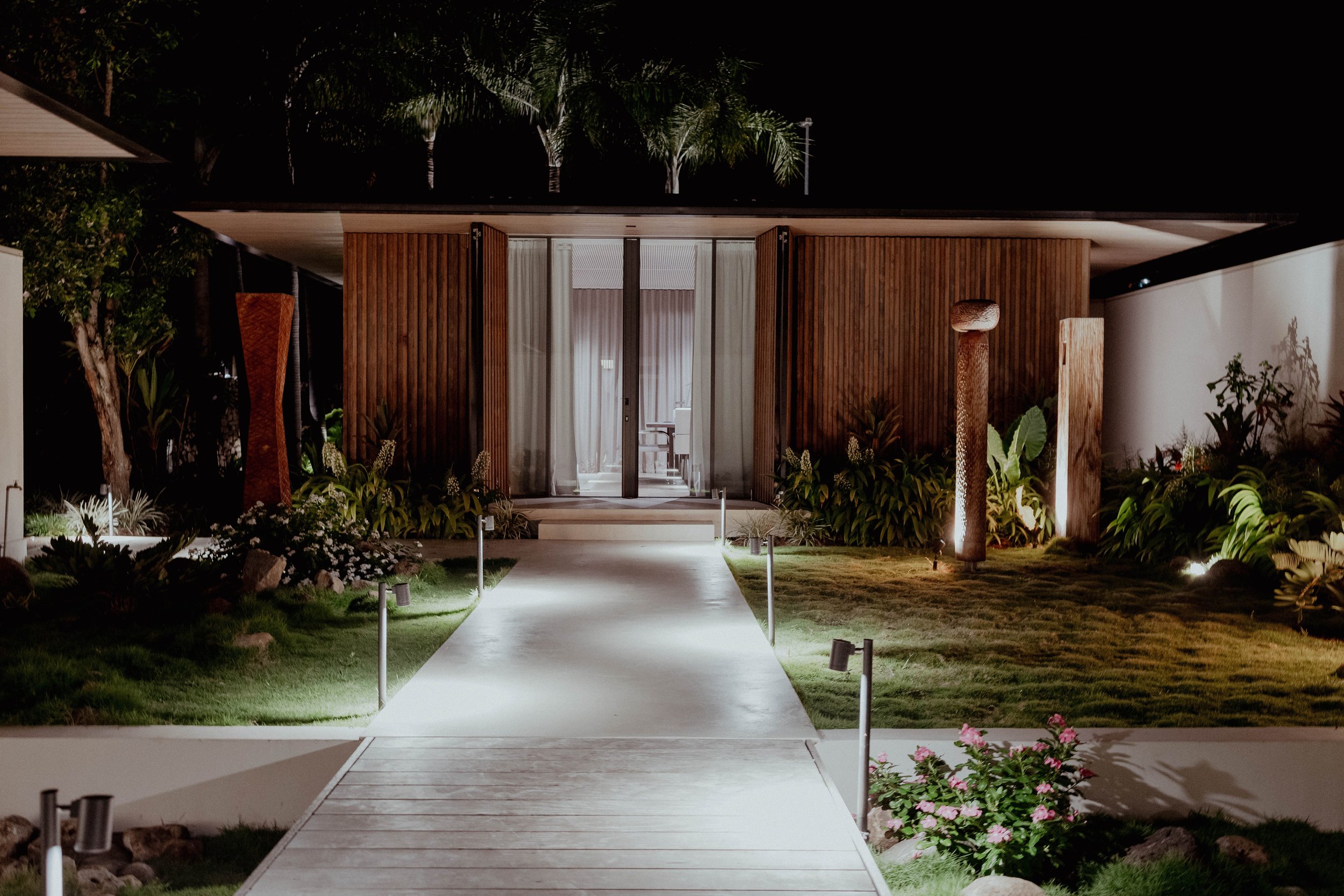 Maison moderne en bois éclairée la nuit, avec un chemin blanc menant à la porte, entourée de végétation et de lampadaires