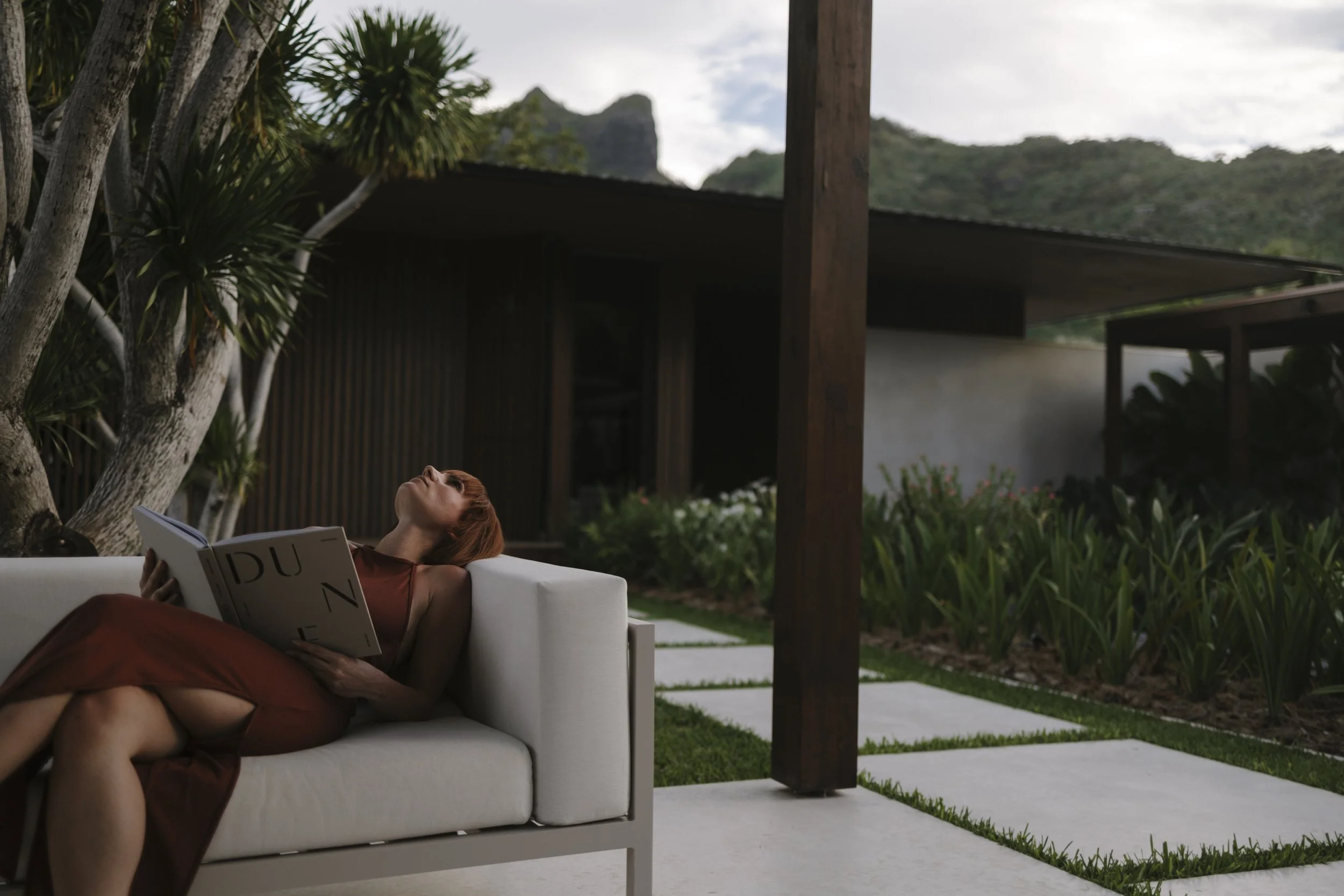 Femme allongée sur un canapé blanc à l'extérieur, lisant un livre, avec un jardin et une maison en arrière-plan.
