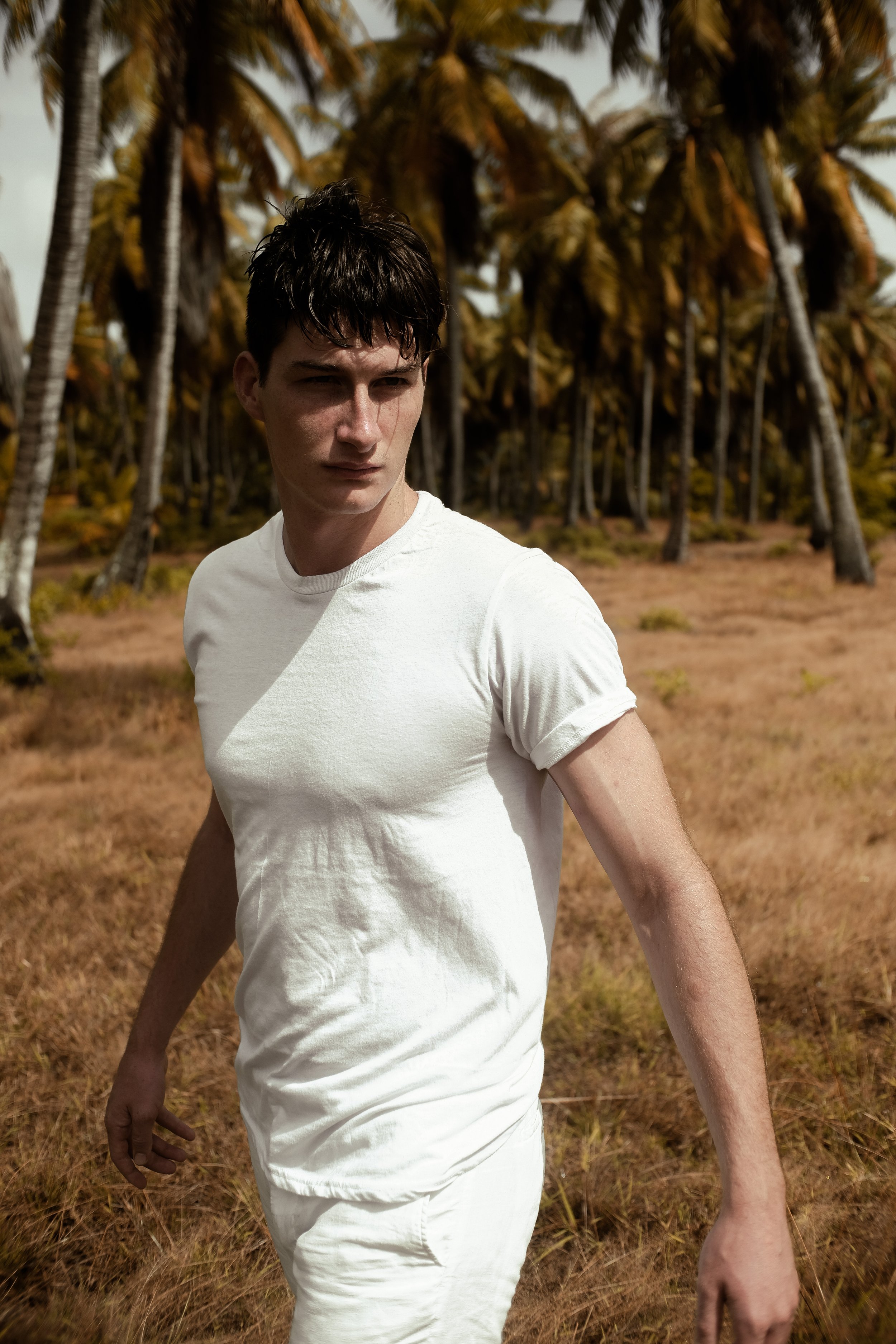 Un jeune homme en t-shirt blanc marche dans une zone herbeuse avec des palmiers en arrière-plan, sous un ciel nuageux.