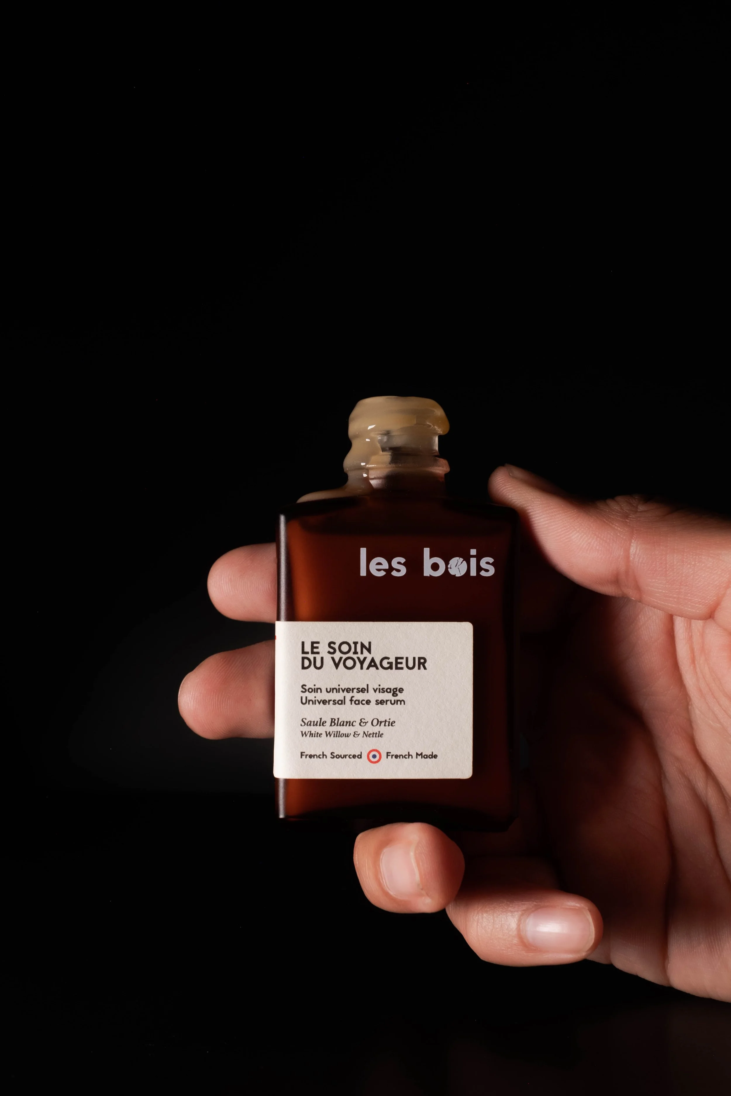 Une main tient un flacon en verre ambré avec une étiquette blanche. Le flacon est étiqueté 'les bois' avec le texte 'LE SOIN DU VOYAGEUR, Soin universel visage, Saule Blanc & Ortie, White Willow & Nettle, French Sourced, French Made'.