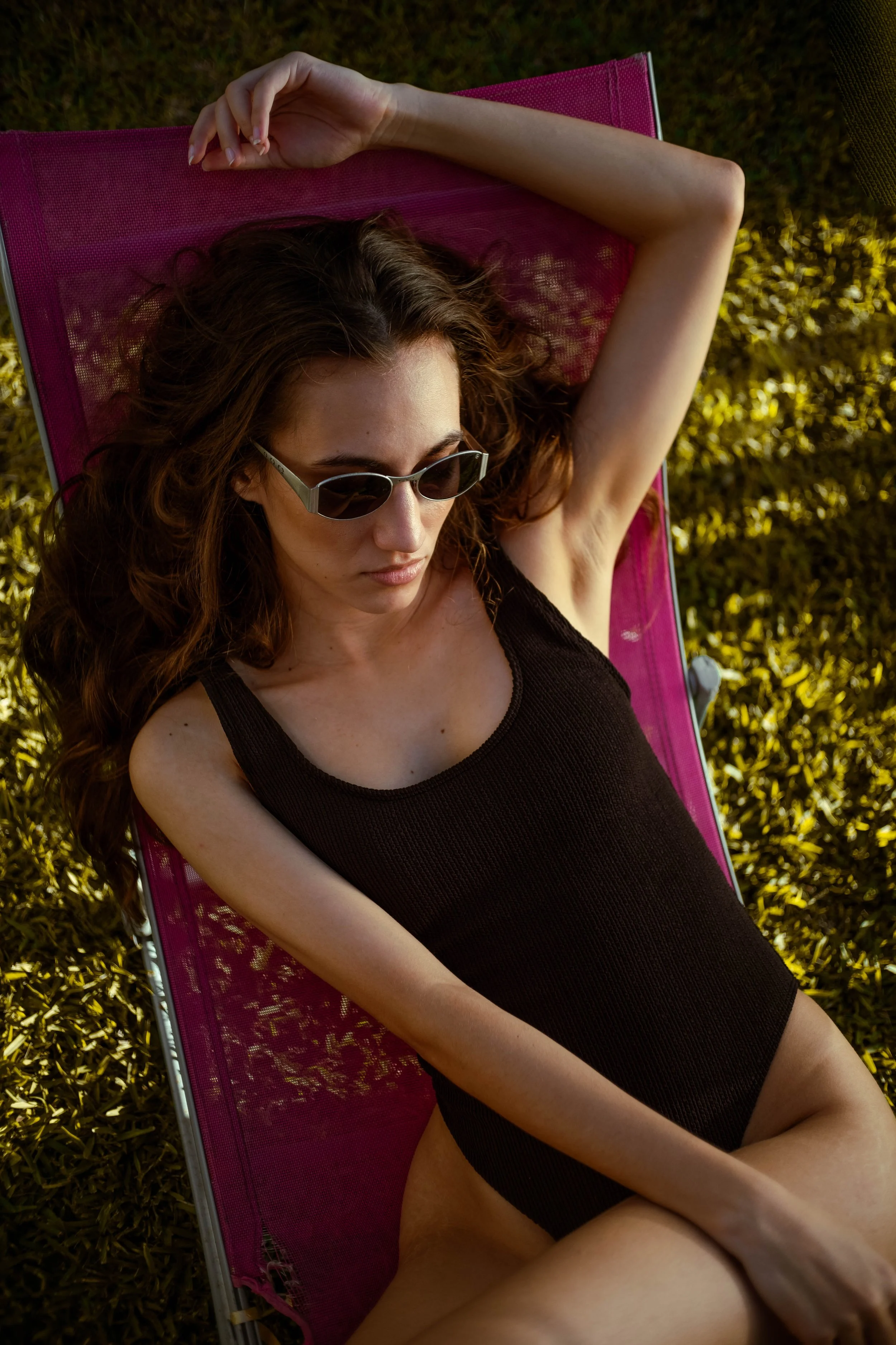 Une femme allongée sur une chaise-pliant rose, portant des lunettes de soleil et un maillot noir, sous la lumière du soleil.