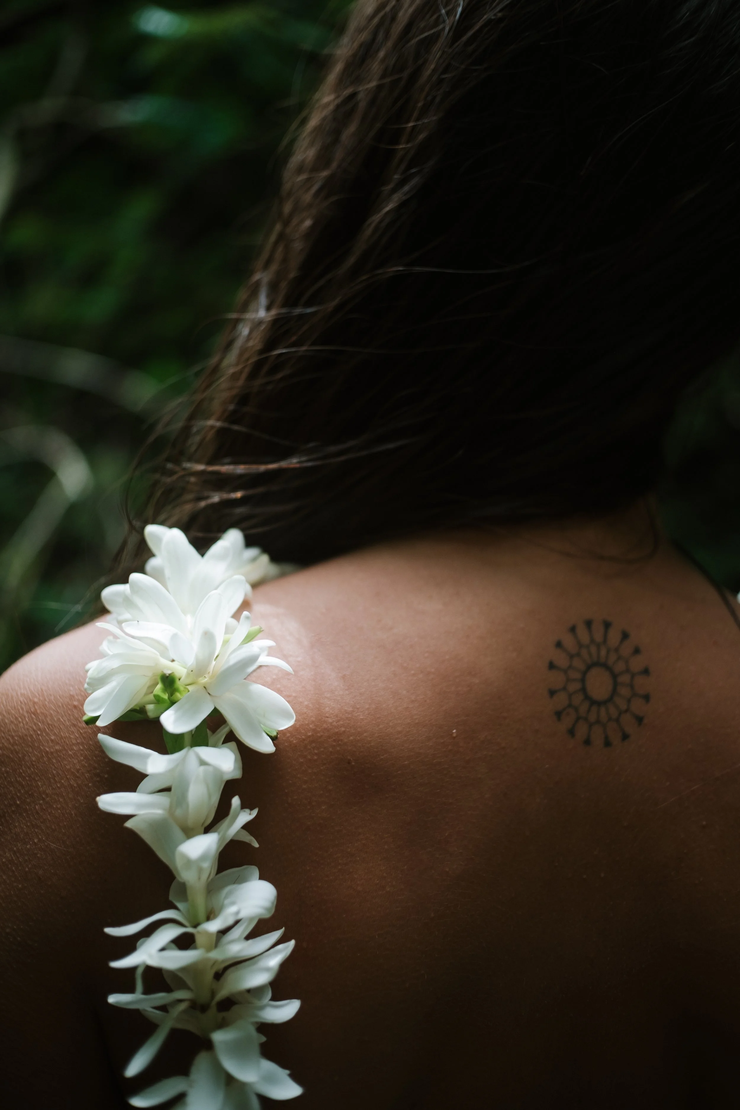 Une épaule d'une personne avec une tatouage ronde et une chaîne de fleurs blanches sur l'épaule, dans un environnement naturel.