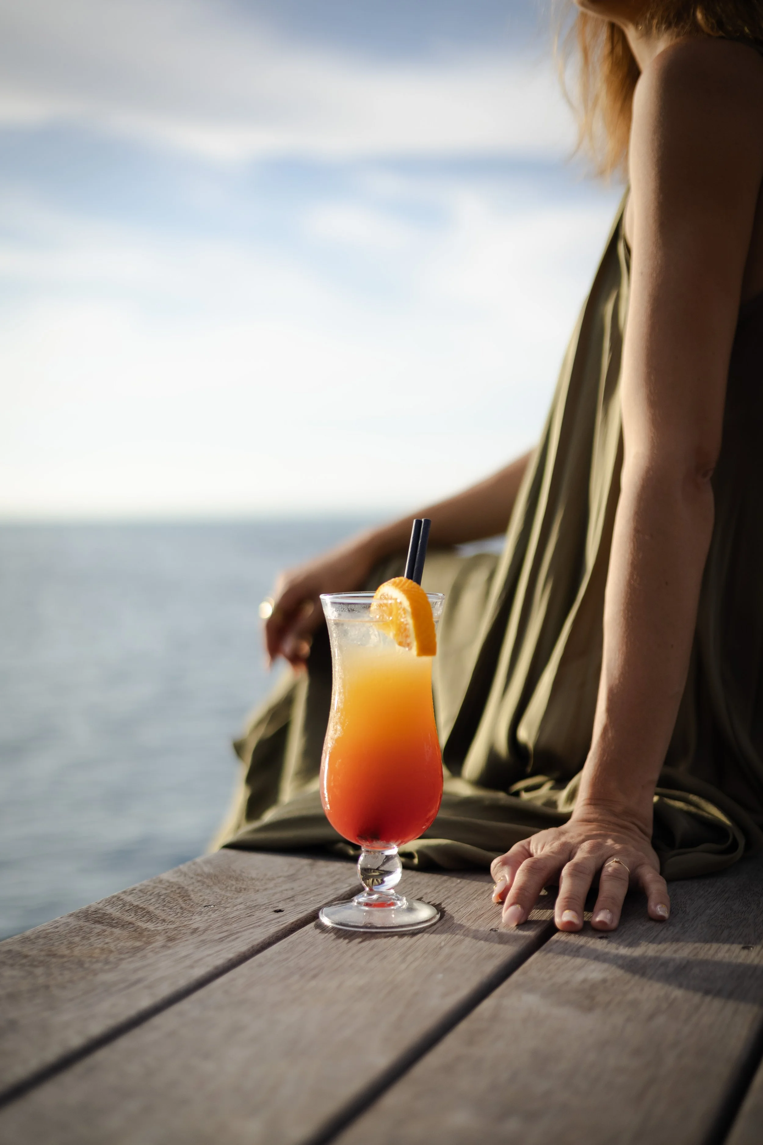 Verre de cocktail coloré avec tranche d'orange et paille, posé sur une table en bois à côté d'une femme assise près de la mer.