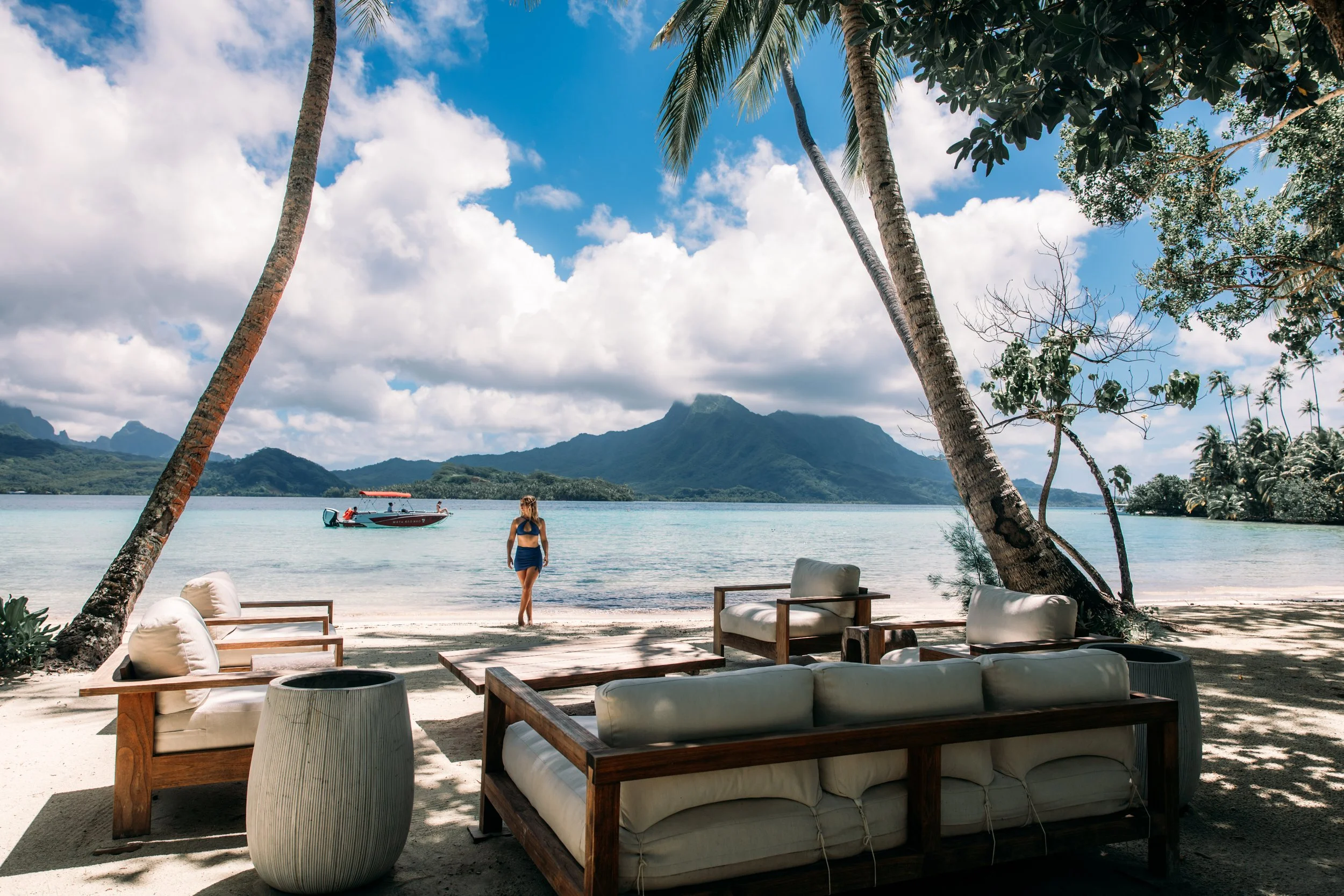 Une plage tropicale avec des palmiers, une femme regardant la mer, un bateau à moteur, des montagnes en arrière-plan et un espace détente avec des canapés en bois et coussins.