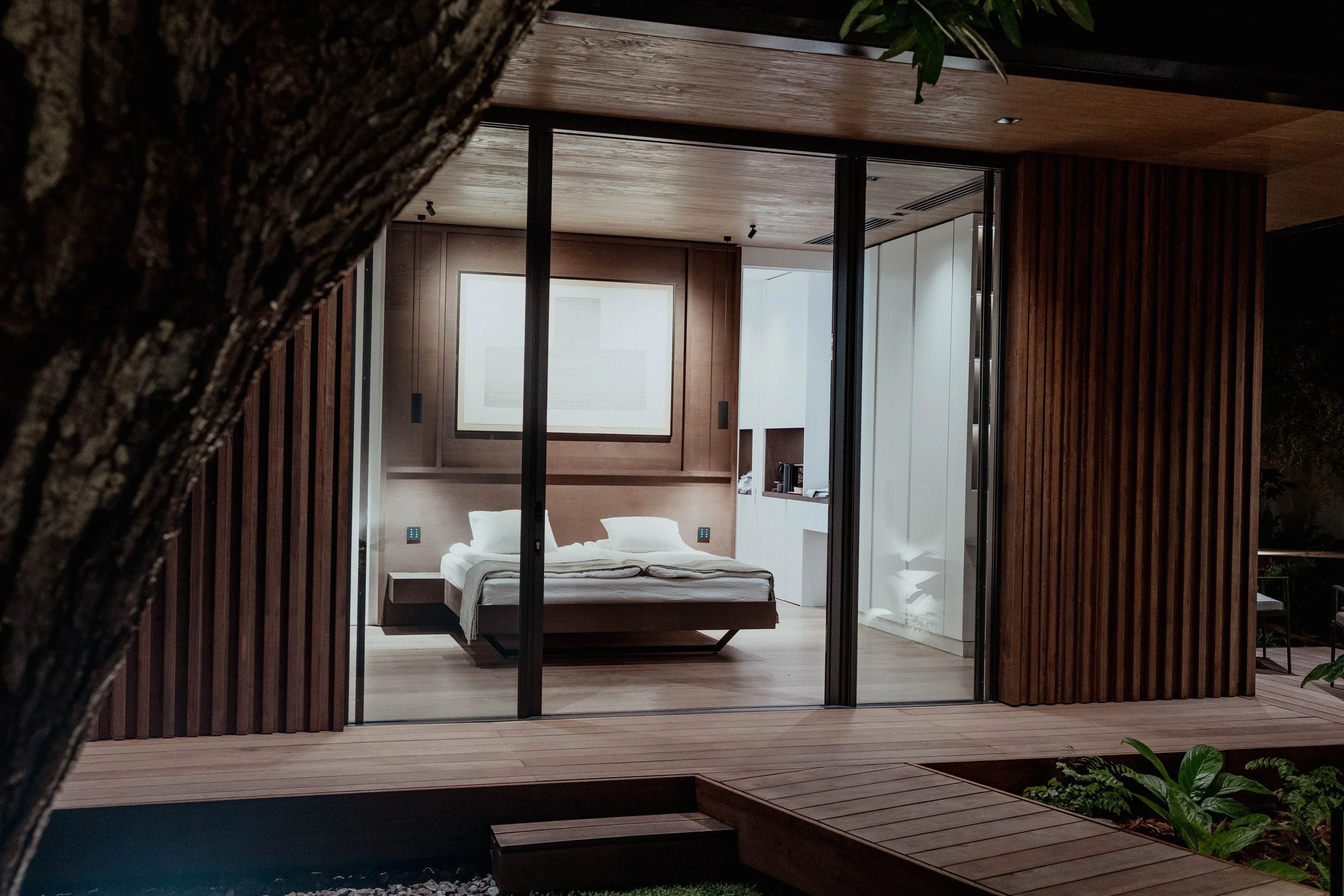 Vue extérieure d'une chambre moderne avec murs en bois, vue à travers une grande baie vitrée vers un lit avec literie blanche à l'intérieur.