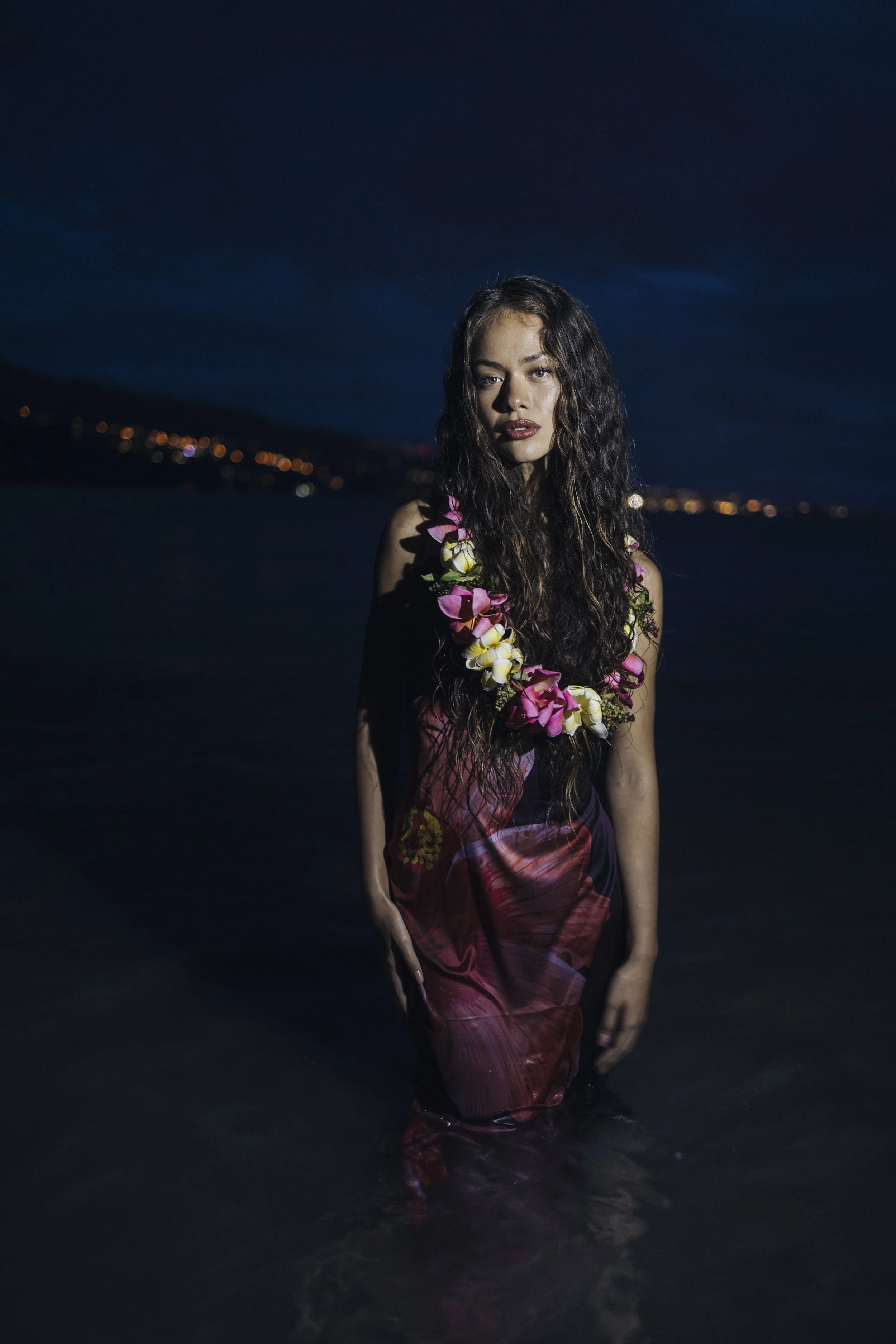 Une femme portant une robe rouge et un collier de fleurs se tient dans la mer la nuit, avec un ciel sombre et des lumières au loin.
