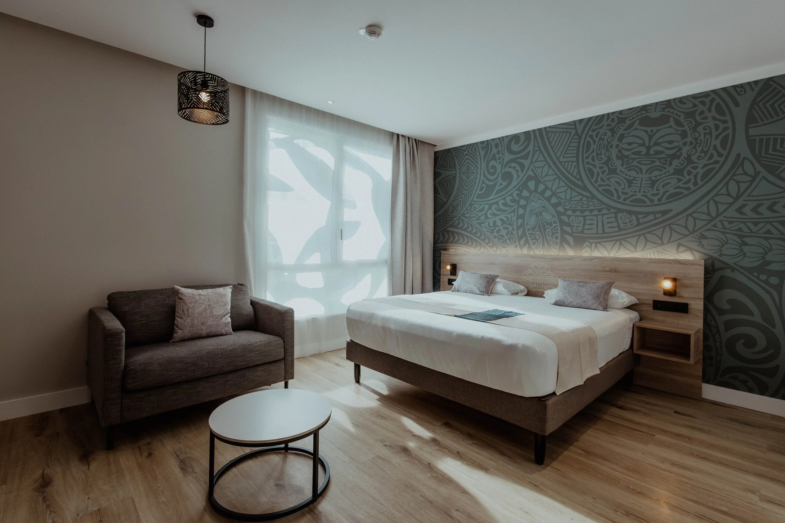 Chambre moderne avec un lit double, un canapé gris, une table ronde, mur d'accent avec un motif tribal, grande fenêtre avec rideaux blancs.