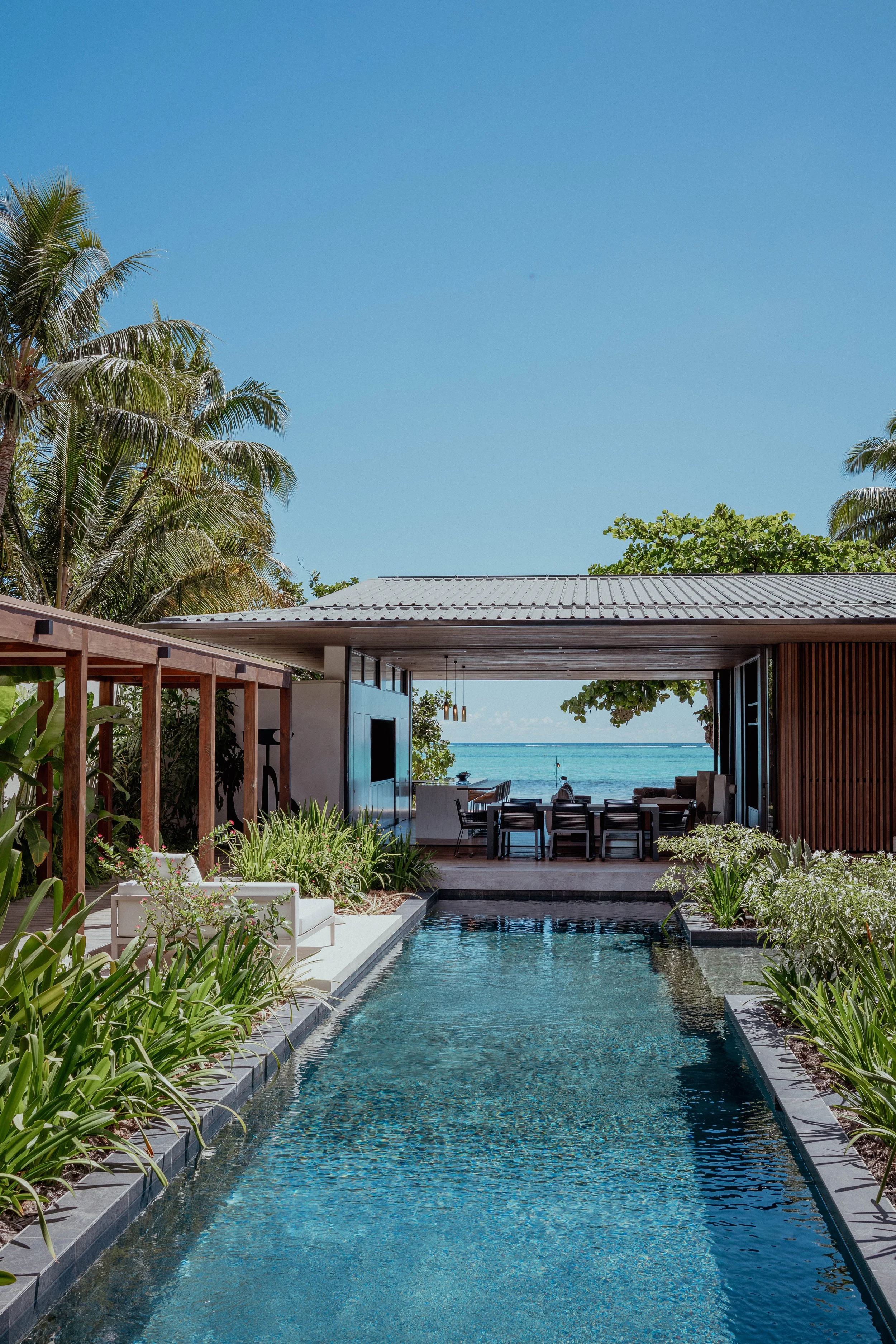 Une villa moderne avec une piscine, un salon ouvert donnant sur la mer, entourée de végétation tropicale, sous un ciel bleu.