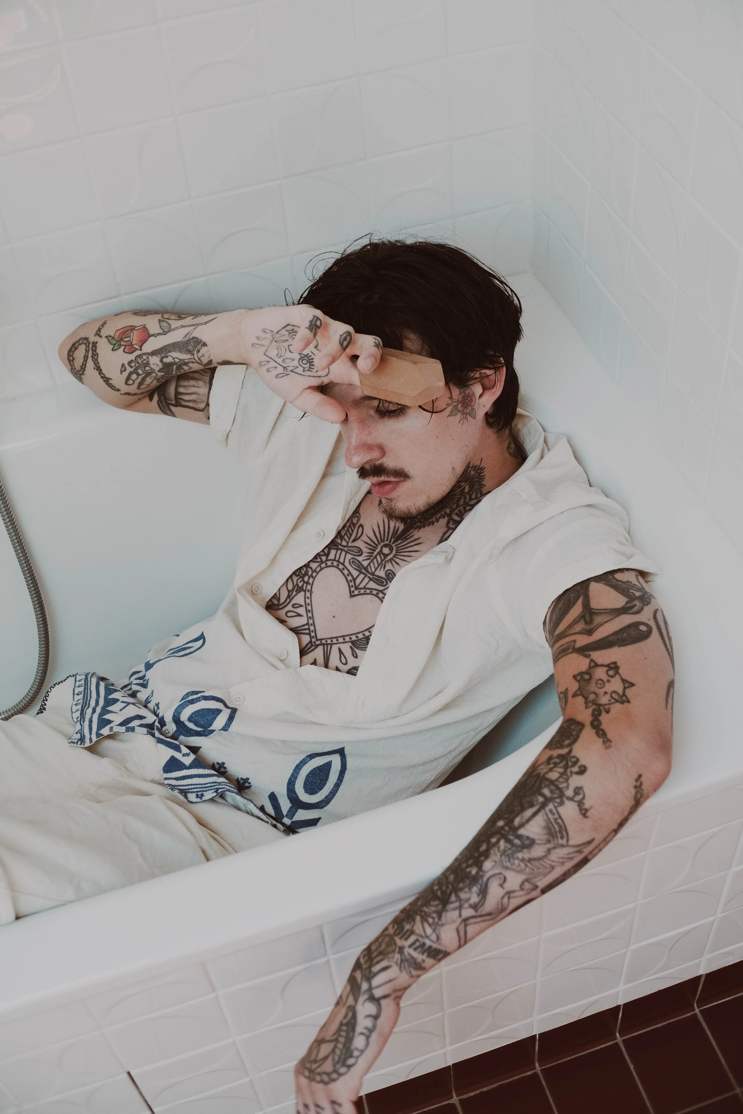 Un homme avec des tatouages allongé dans une baignoire, l'air fatigué, tenant un objet en pierre sur son front. Tattoos visibles sur ses bras et son torse, portant une chemise blanche ouverte.