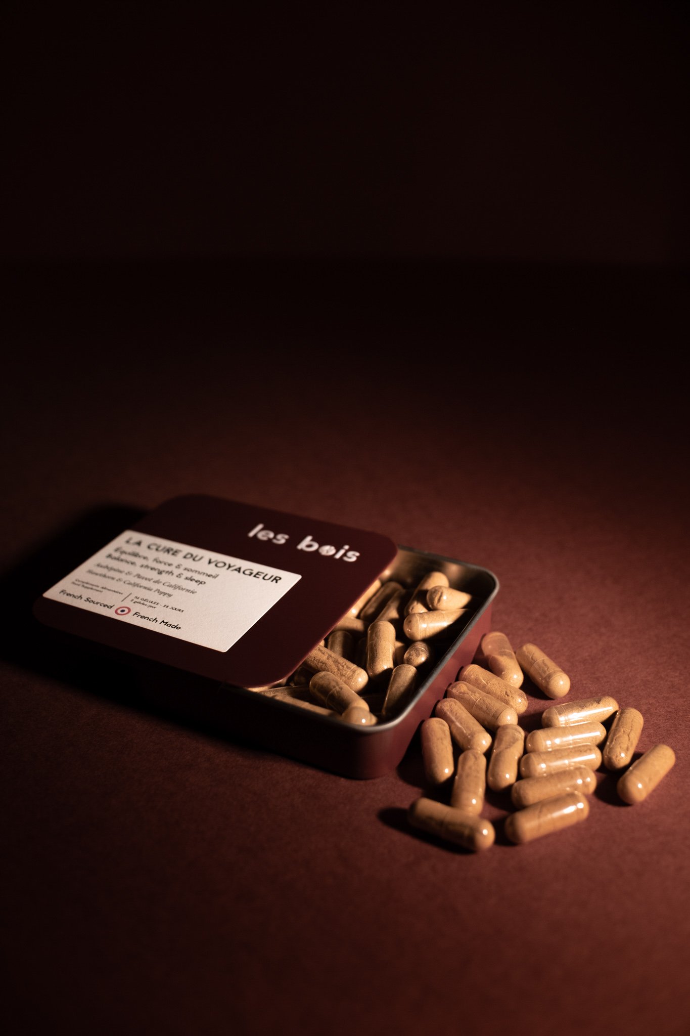 Une boîte metalique ouverte contenant des capsules de médicaments, avec quelques capsules qui se sont échappées sur une surface en bois et un fond sombre.