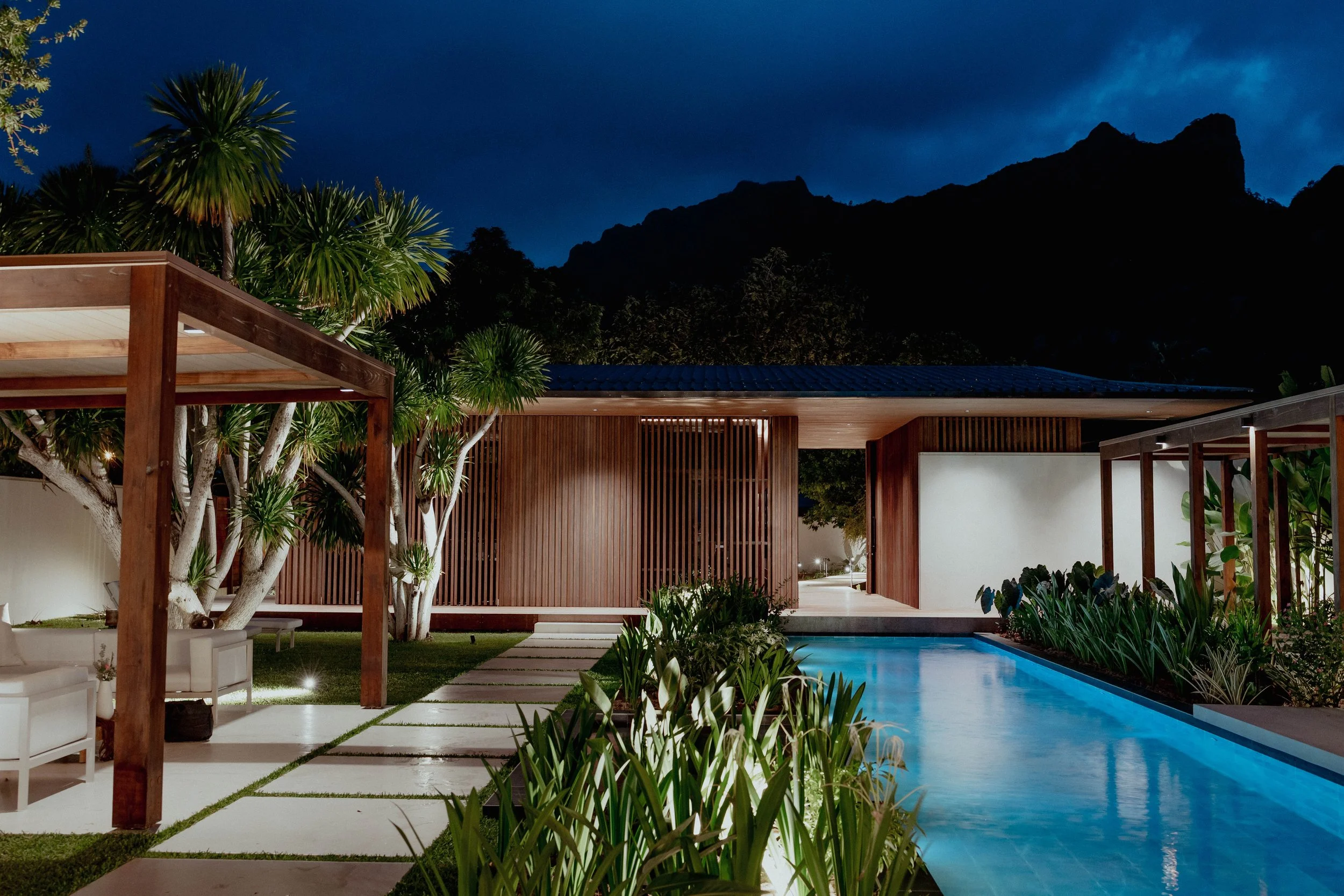 Vue nocturne d'une piscine illuminée entourée de végétation et d'une architecture moderne en bois, avec un ciel sombre en arrière-plan.
