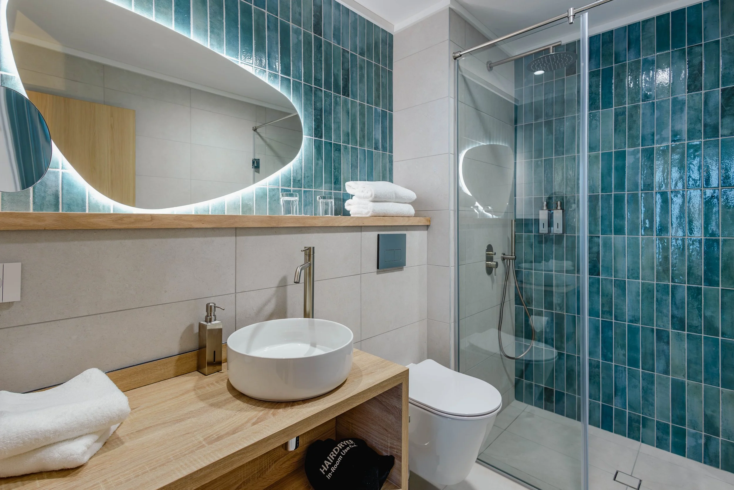 Salle de bain moderne avec miroir éclairé, lavabo en acier inoxydable sur un meuble en bois, toilettes, serviettes blanches, douche en verre avec céramiques bleues murales.