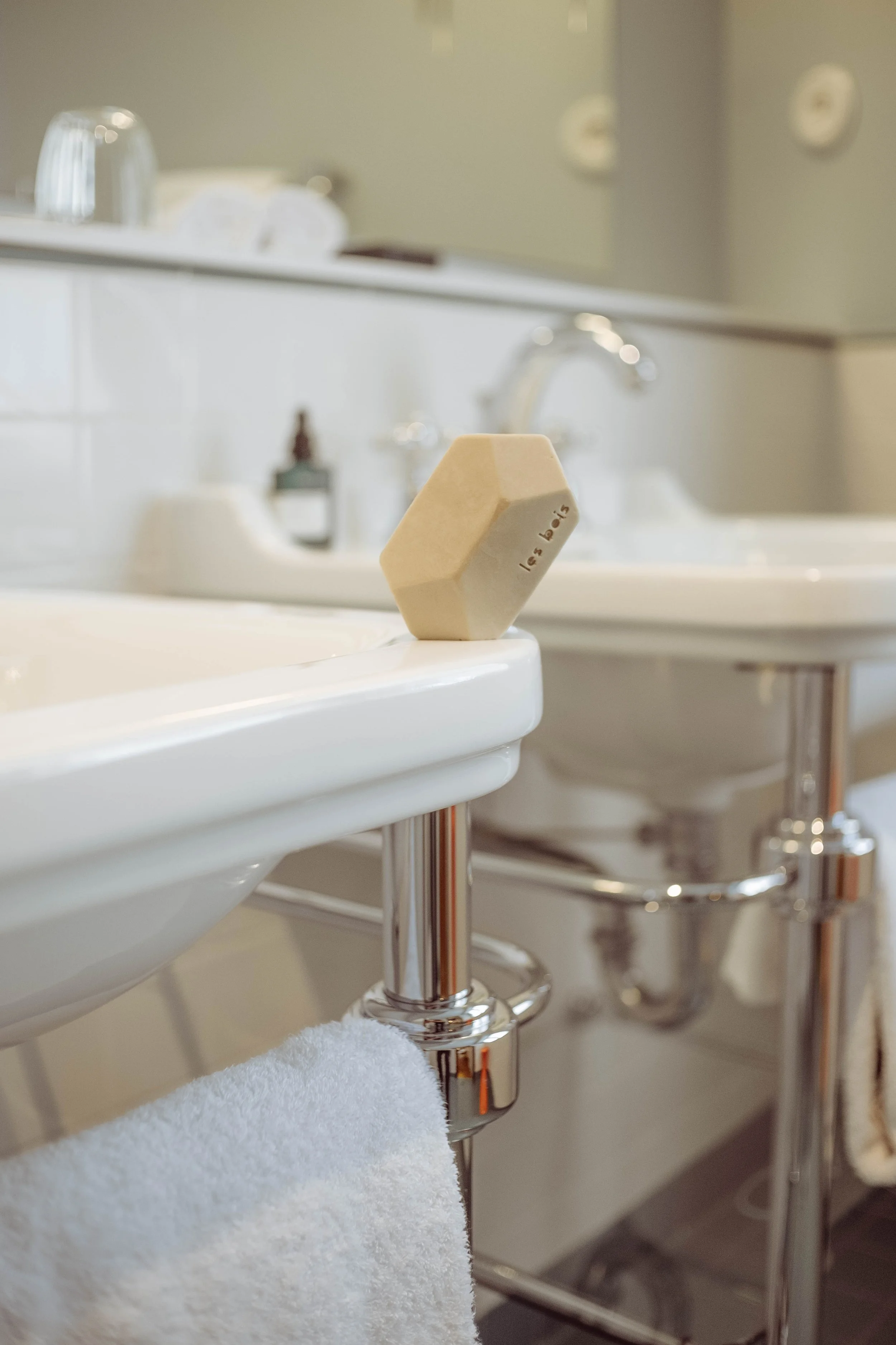 Un savon beige posé sur le rebord d'un lavabo blanc dans une salle de bains. En arrière-plan, on aperçoit un miroir, un robinet et d'autres éléments de salle de bains.