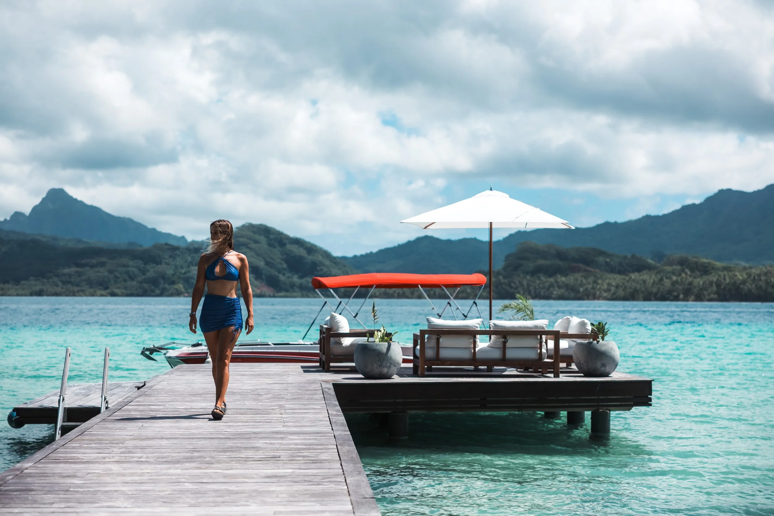 Femme en maillot de bain sur un pont en bois au bord de l'eau turquoise avec des montagnes en arrière-plan, un bateau, un parasol blanc, un canapé et des pots de fleurs sur une plateforme flottante.