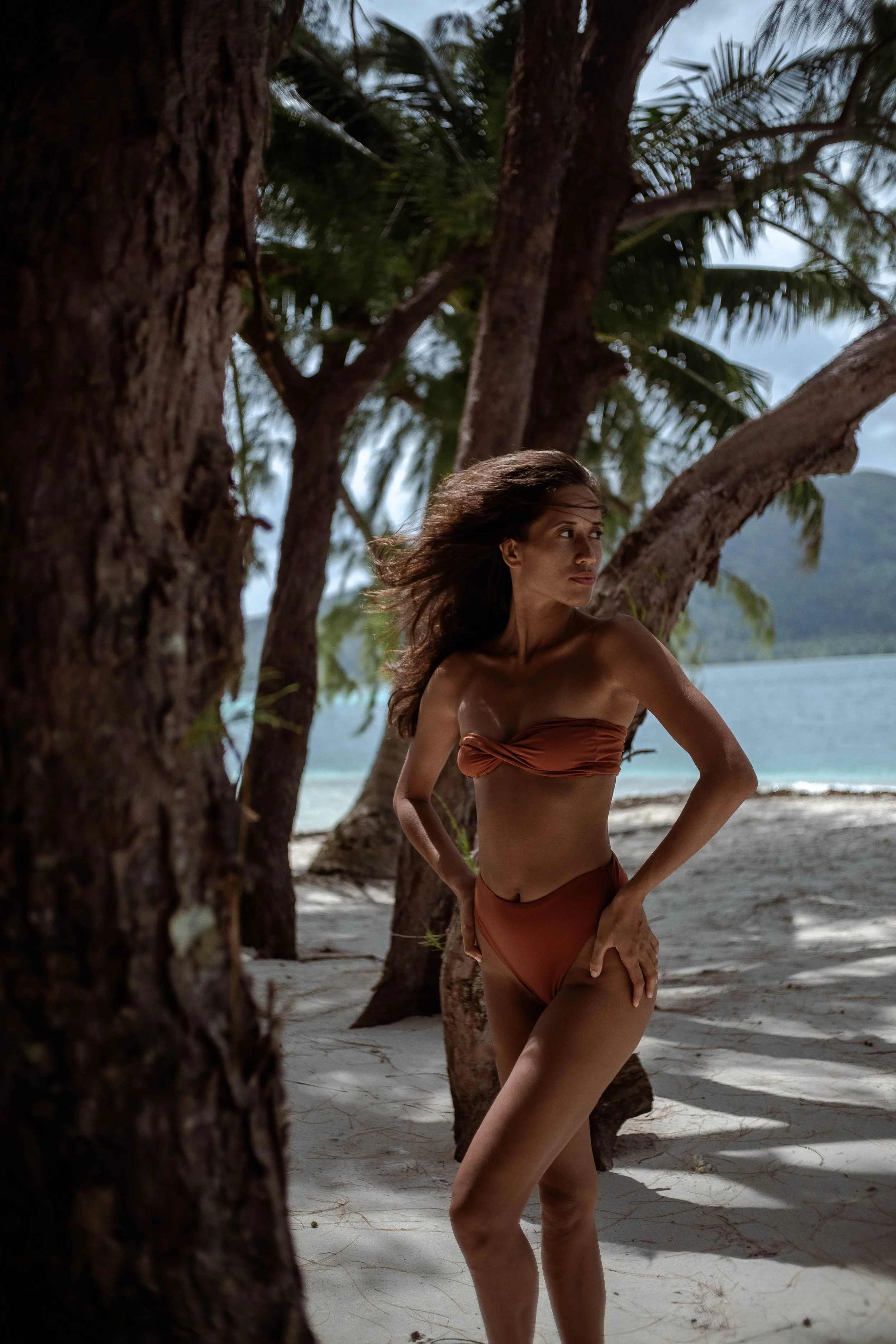 Femme en maillot de bain sur une plage avec des arbres et la mer en arrière-plan.