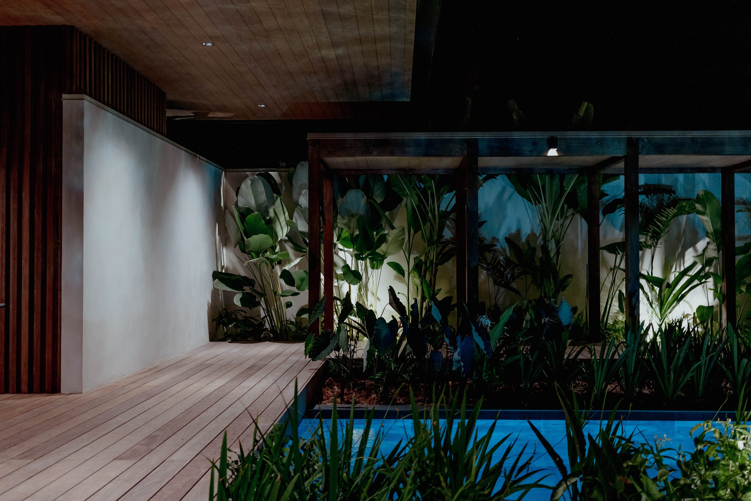 Un patio moderne avec une terrasse en bois, des plantes vertes, une piscine et une structure en bois, éclairé la nuit.