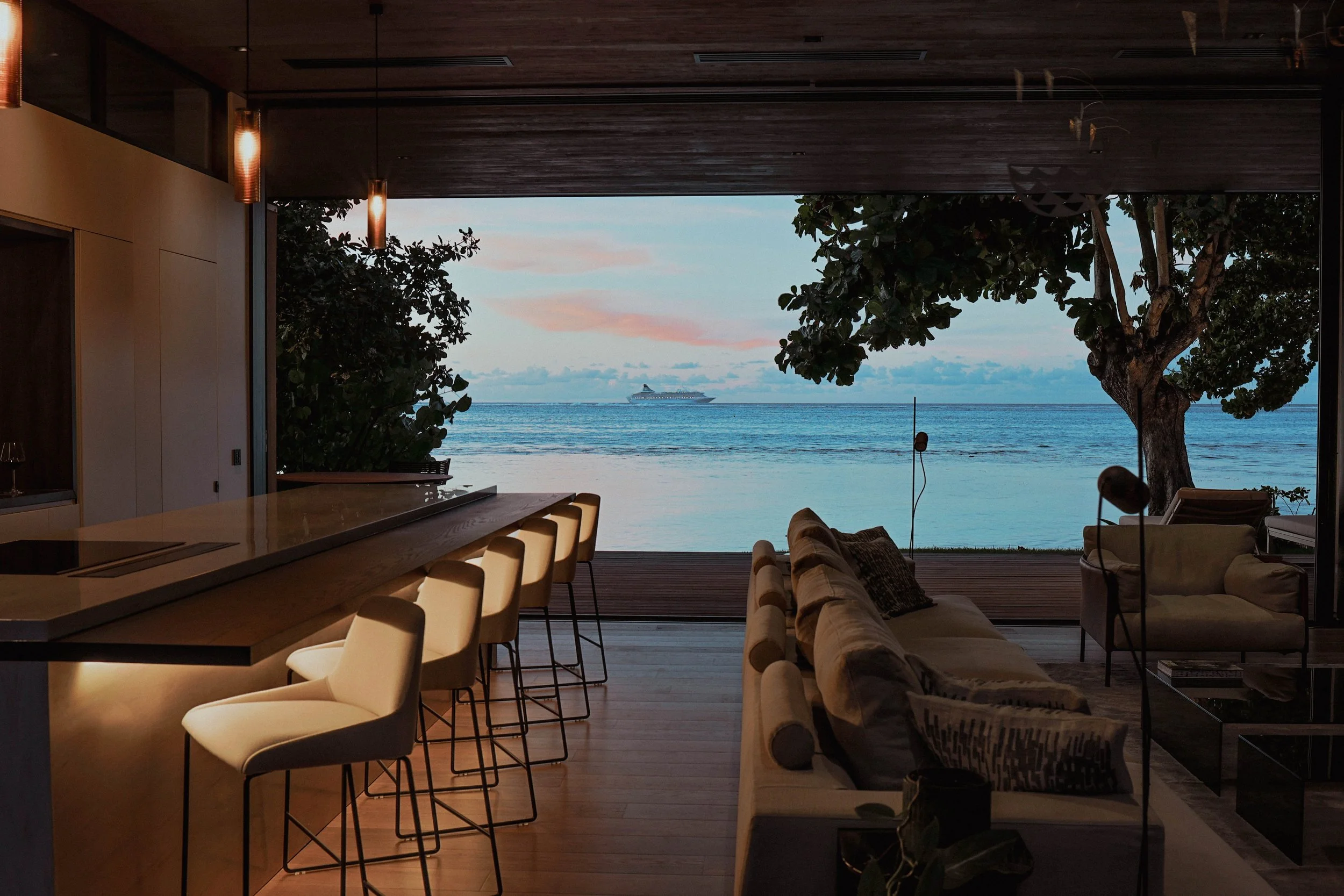 Salon intérieur avec vue sur la mer, canapés, table basse, et arbres, avec un bateau en mer au loin, lors du coucher du soleil.