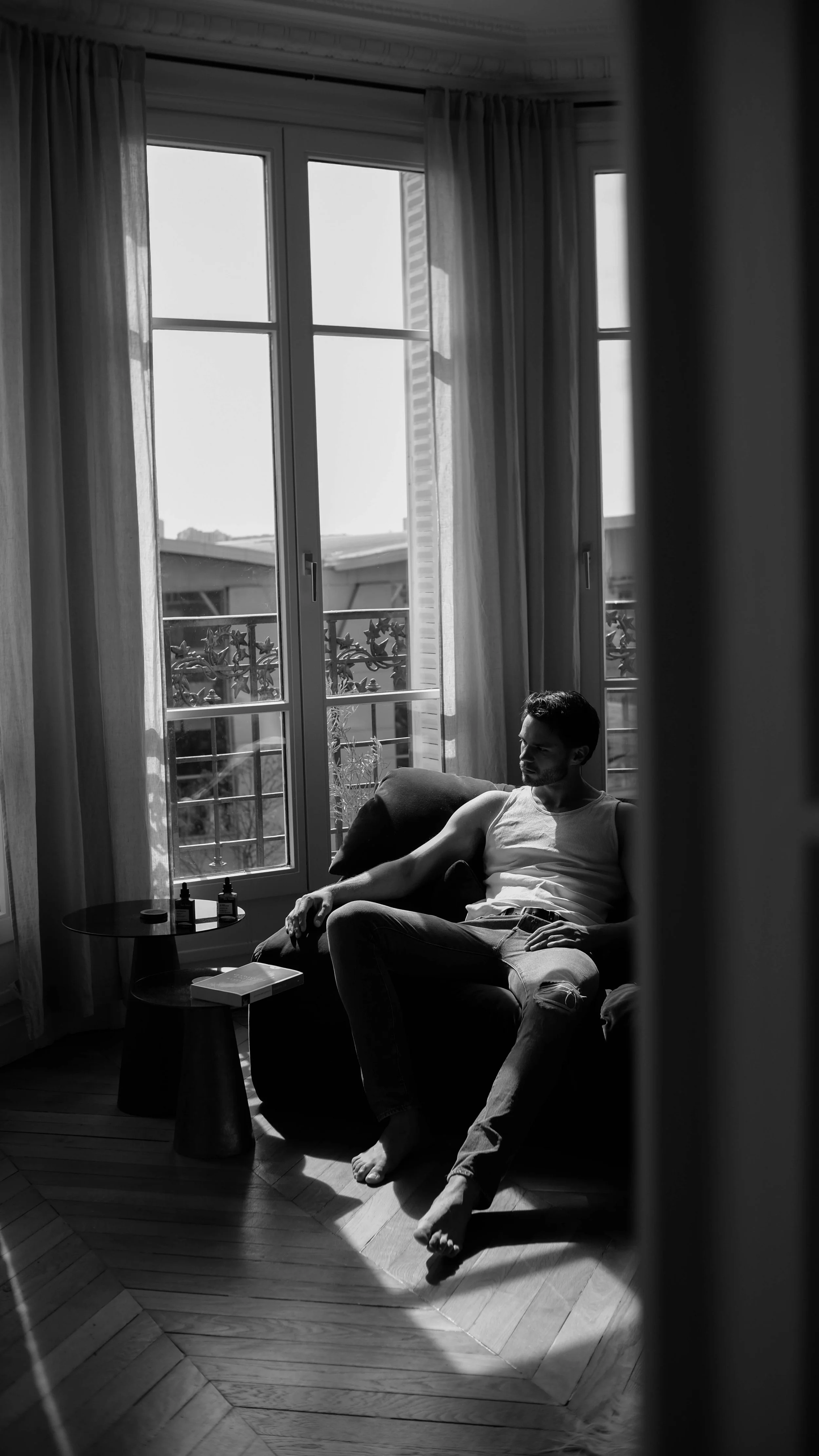 Un homme assis sur un canapé dans une pièce lumineuse, avec de grandes fenêtres laissant entrer la lumière du jour, en noir et blanc.
