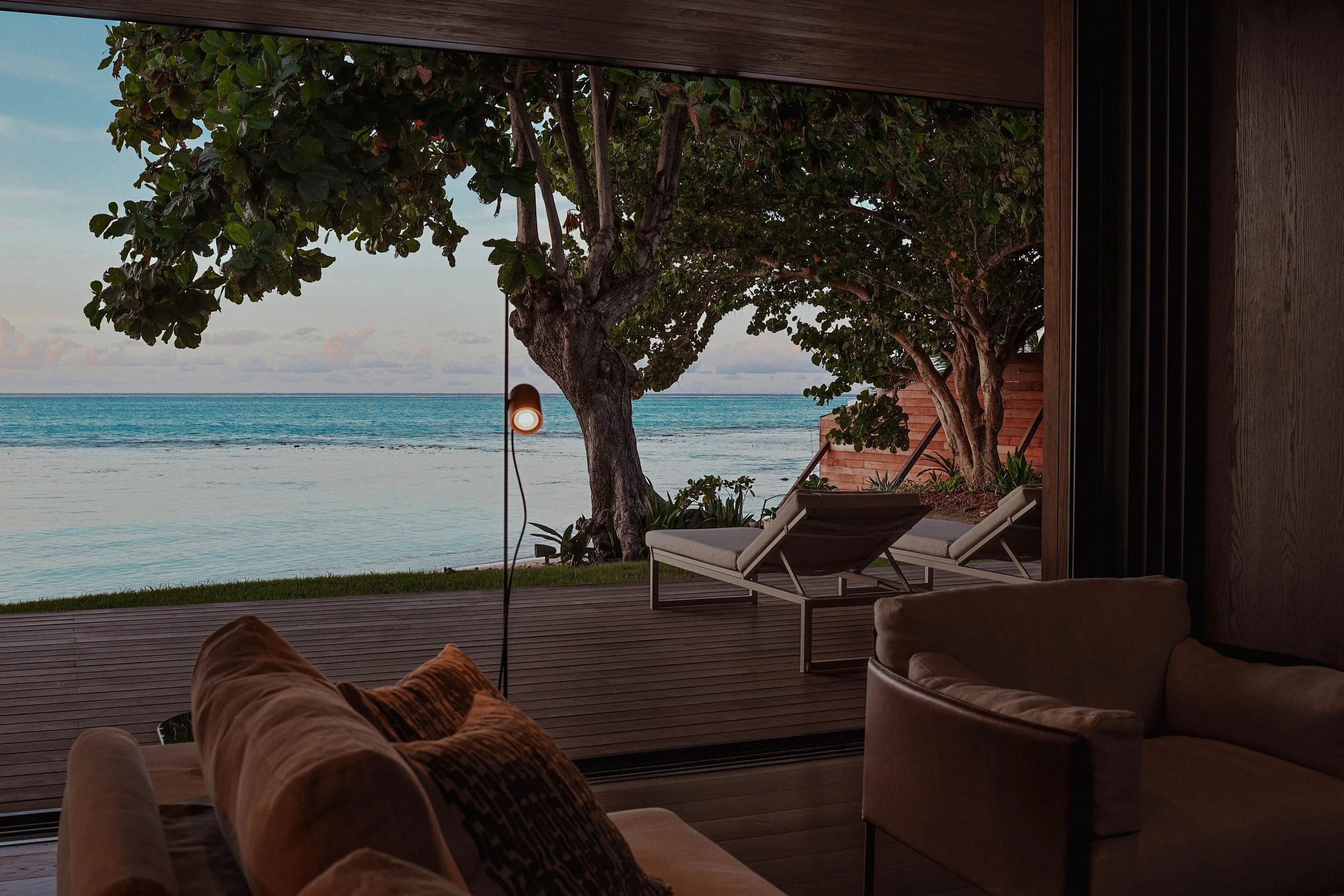 Intérieur d'une maison avec un grand mur en verre ouvrant sur une terrasse en bois face à la mer, avec des arbres et des chaises longues.