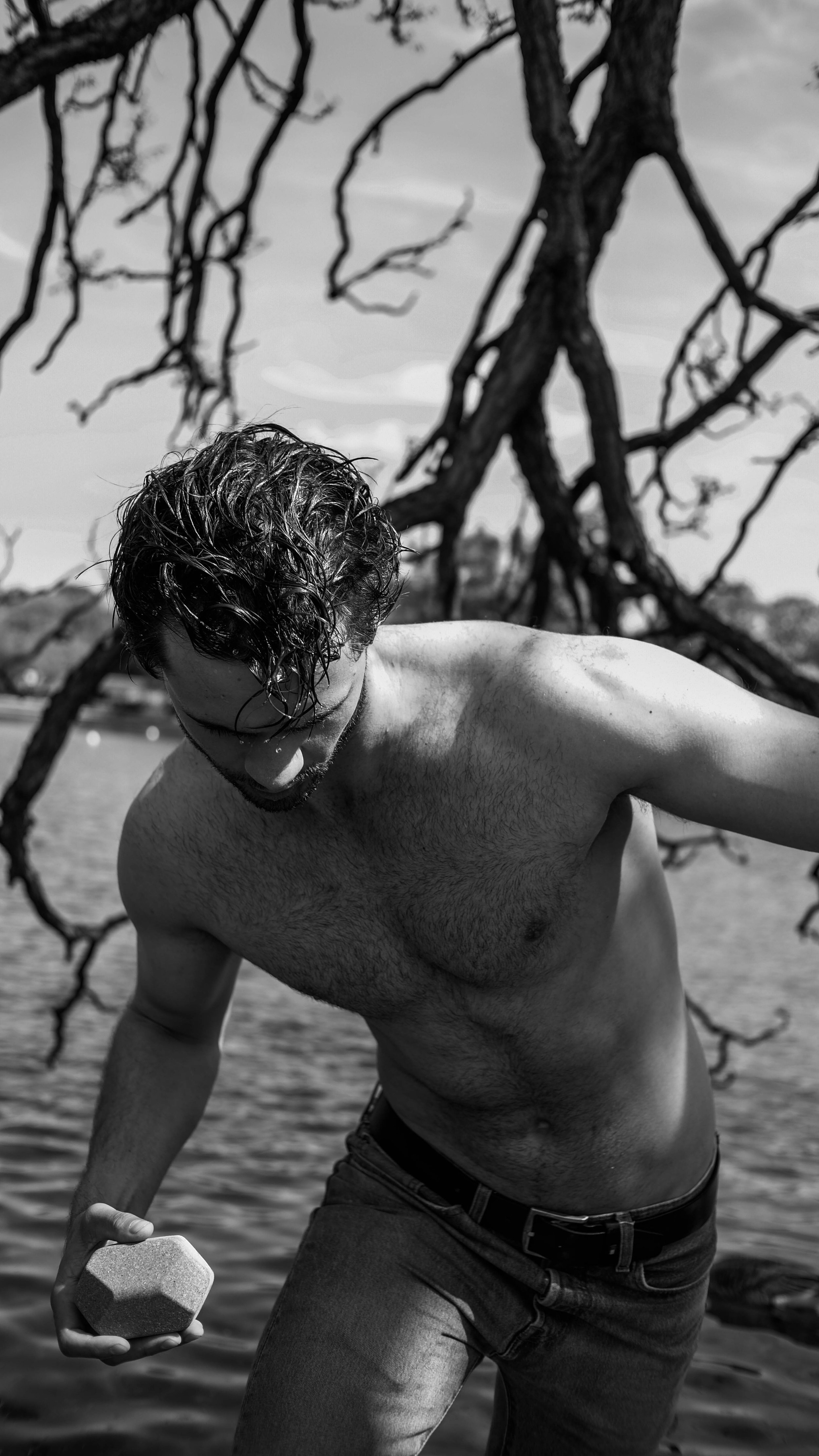 Jeune homme barbu et moustachu, sans chemise, tenant une pierre, debout près d'un lac avec un arbre sans feuilles en arrière-plan, en noir et blanc.