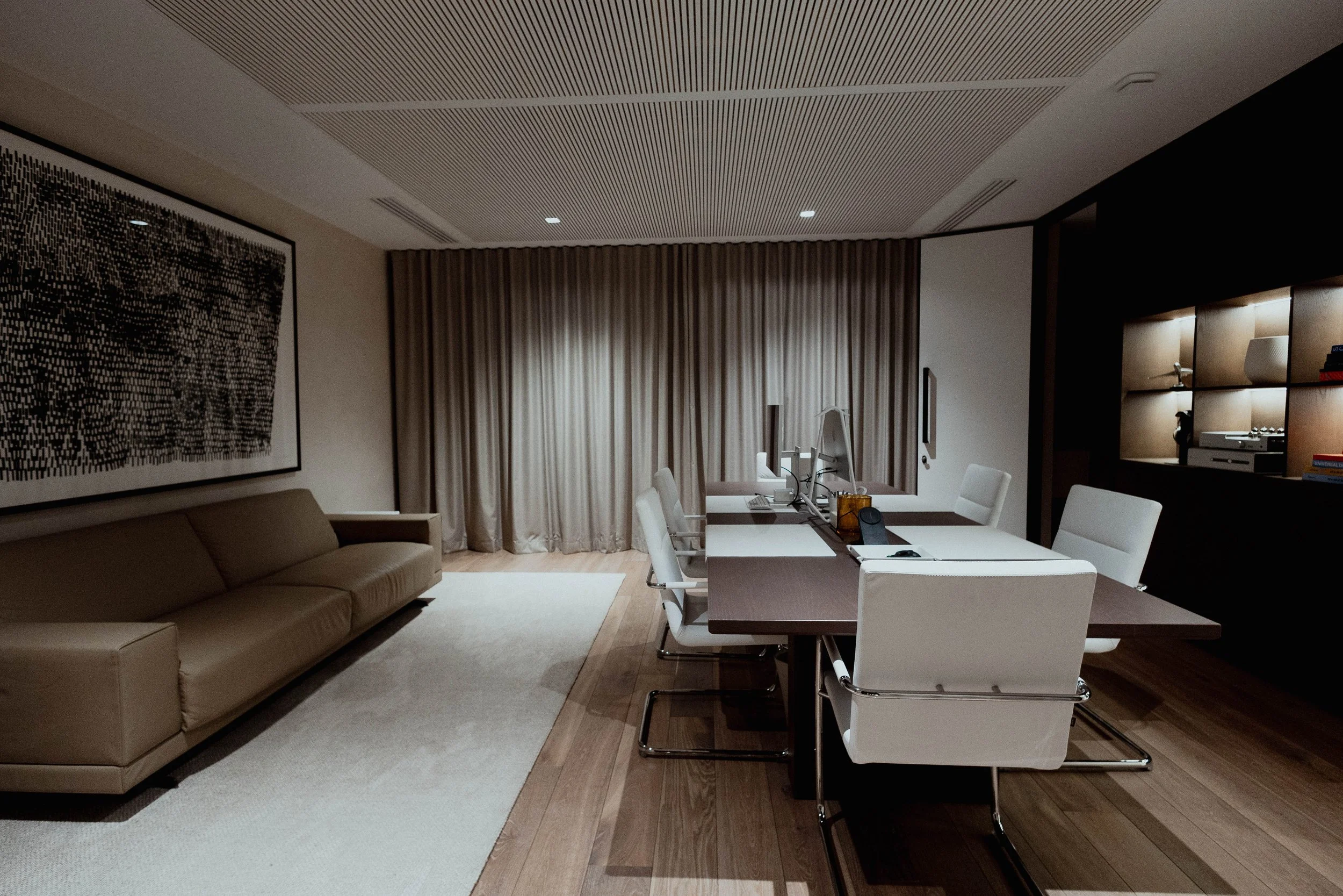 Salle de réunion moderne avec table, chaises blanches, canapé beige, grande œuvre d'art noire et blanche, étagère avec livres et lampes, murs modernes, sol en bois clair, rideaux beige.