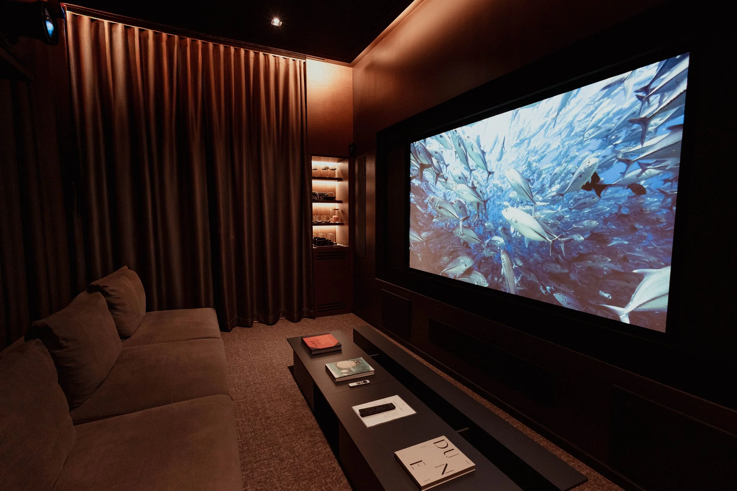Une salle de cinéma sombre avec un grand écran montrant une scène sous-marine avec un banc de poissons. Il y a un canapé beige à gauche, une table basse avec des livres, un téléphone portable et un magazine. Des étagères avec des objets sont visibles