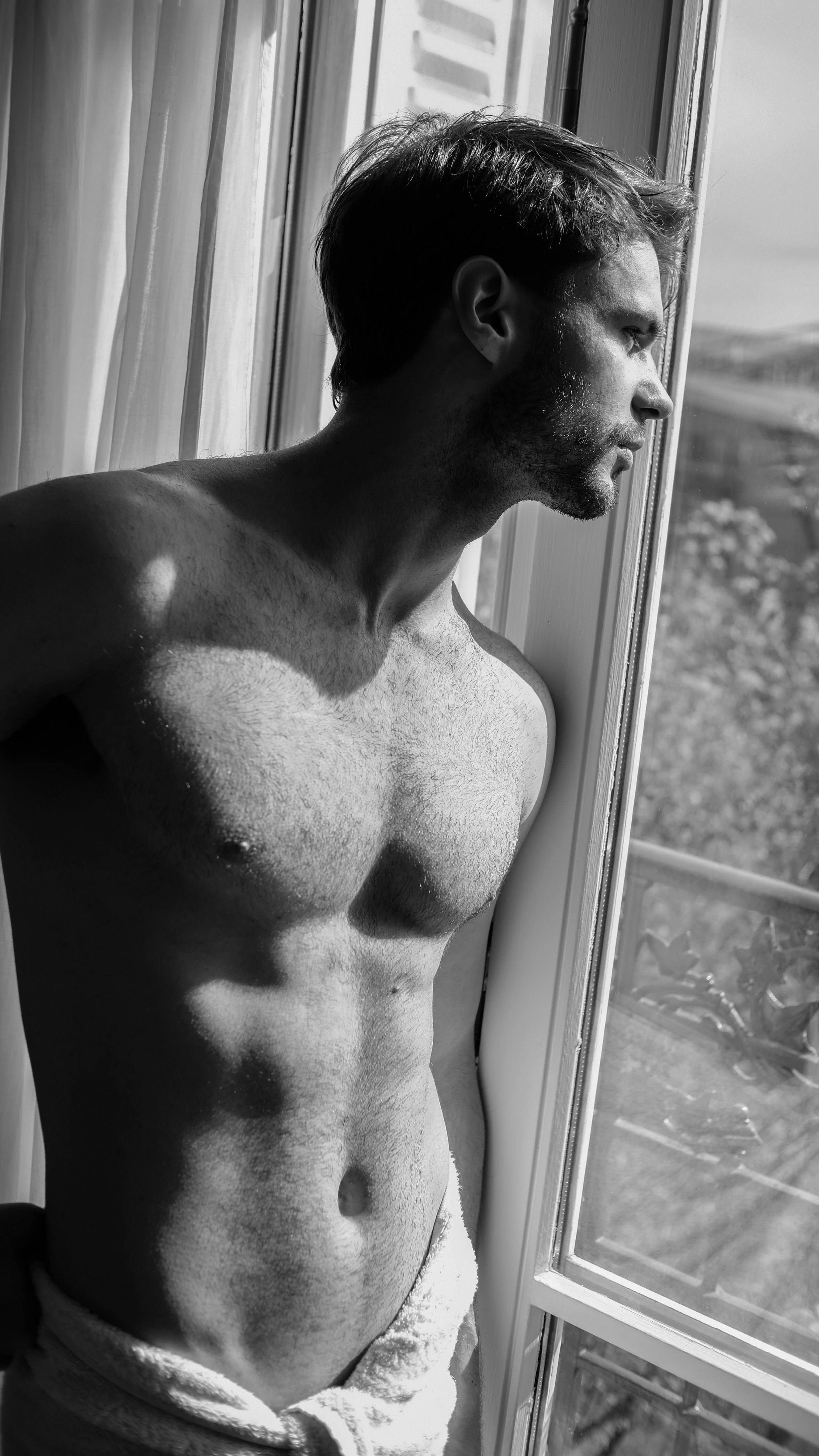 Homme debout près d'une fenêtre, regardant dehors, en noir et blanc, torse nu, avec des cheveux courts et une barbe, laissant entrer la lumière naturelle.