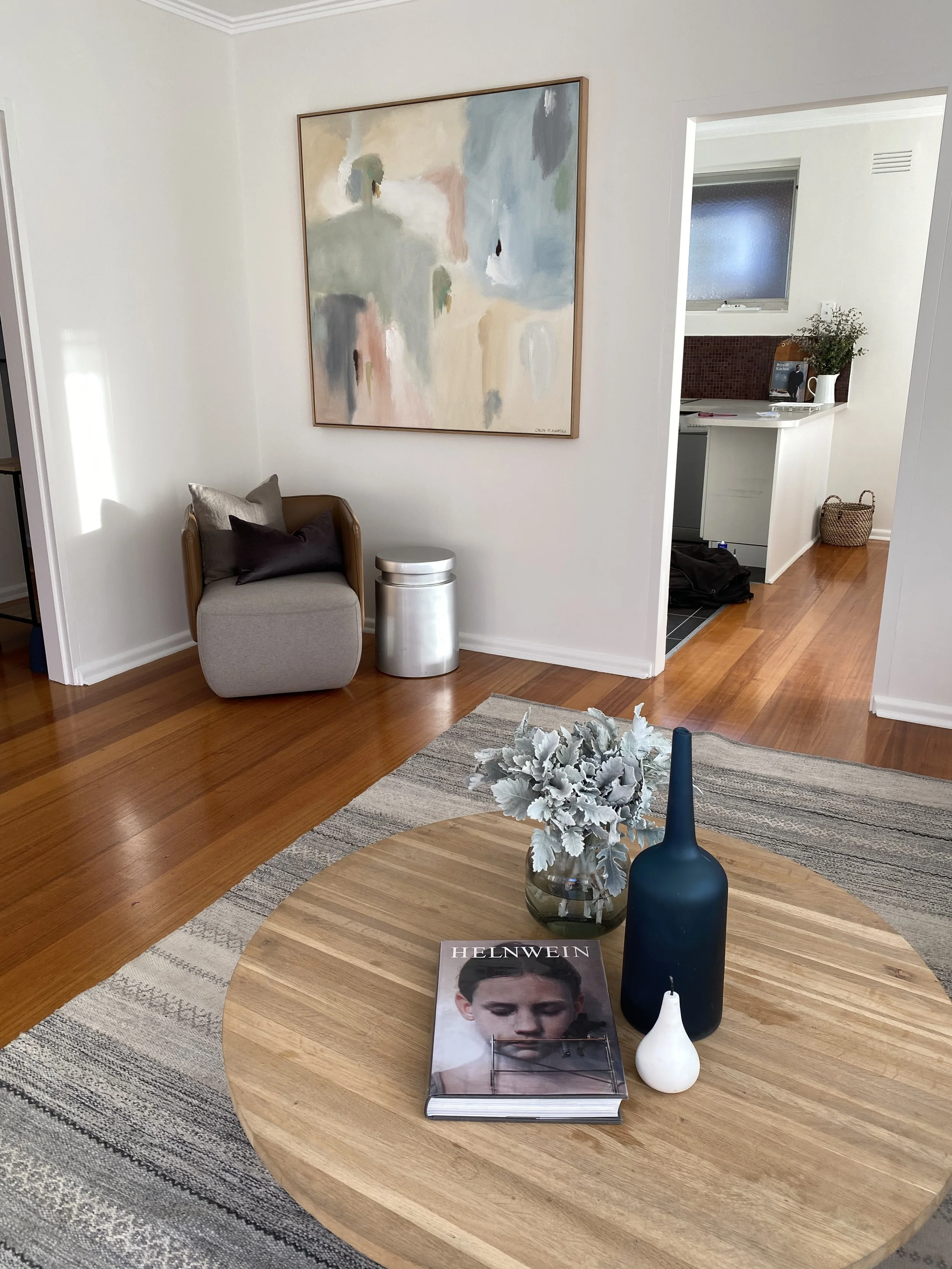 A cozy living room corner featuring a round wooden table with a blue vase, a white decorative item, a gray and white flower arrangement, and a magazine. In the background, there is a gray armchair with pillows, a metallic side table, an abstract pain