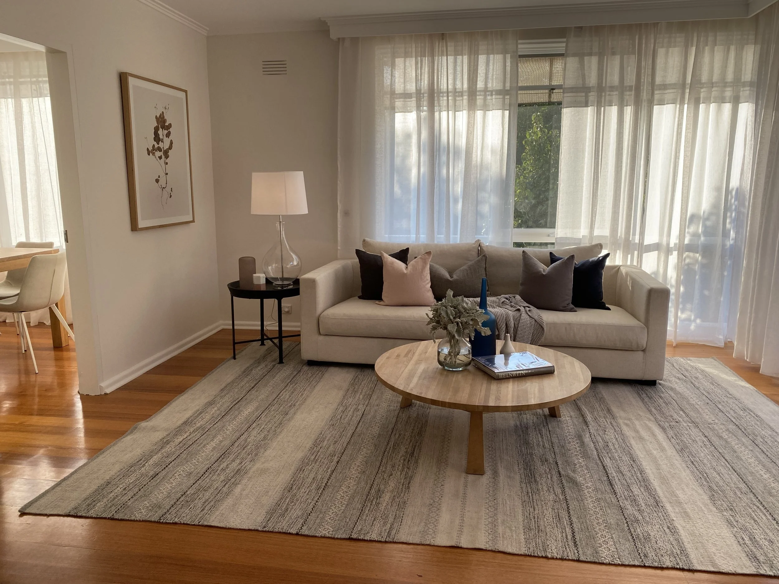 Living room with cream sofa, dark and light throw pillows, round wooden coffee table with decorative vases, book, and plant, sheer curtains, wooden floor, side table with lamp, framed artwork on wall.