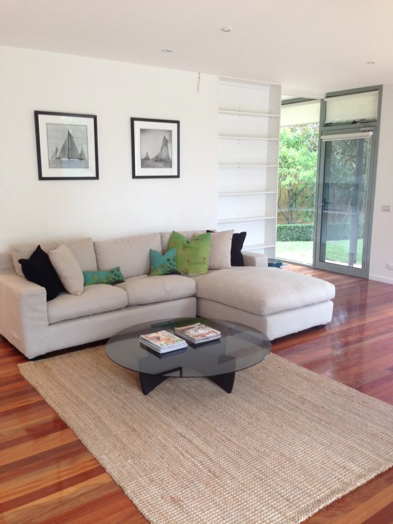 Living room with a beige sectional sofa with black and green throw pillows, a black round coffee table with magazines, a beige area rug, and two black-and-white framed sailboat pictures on the wall. A large window with a garden view and white shelves