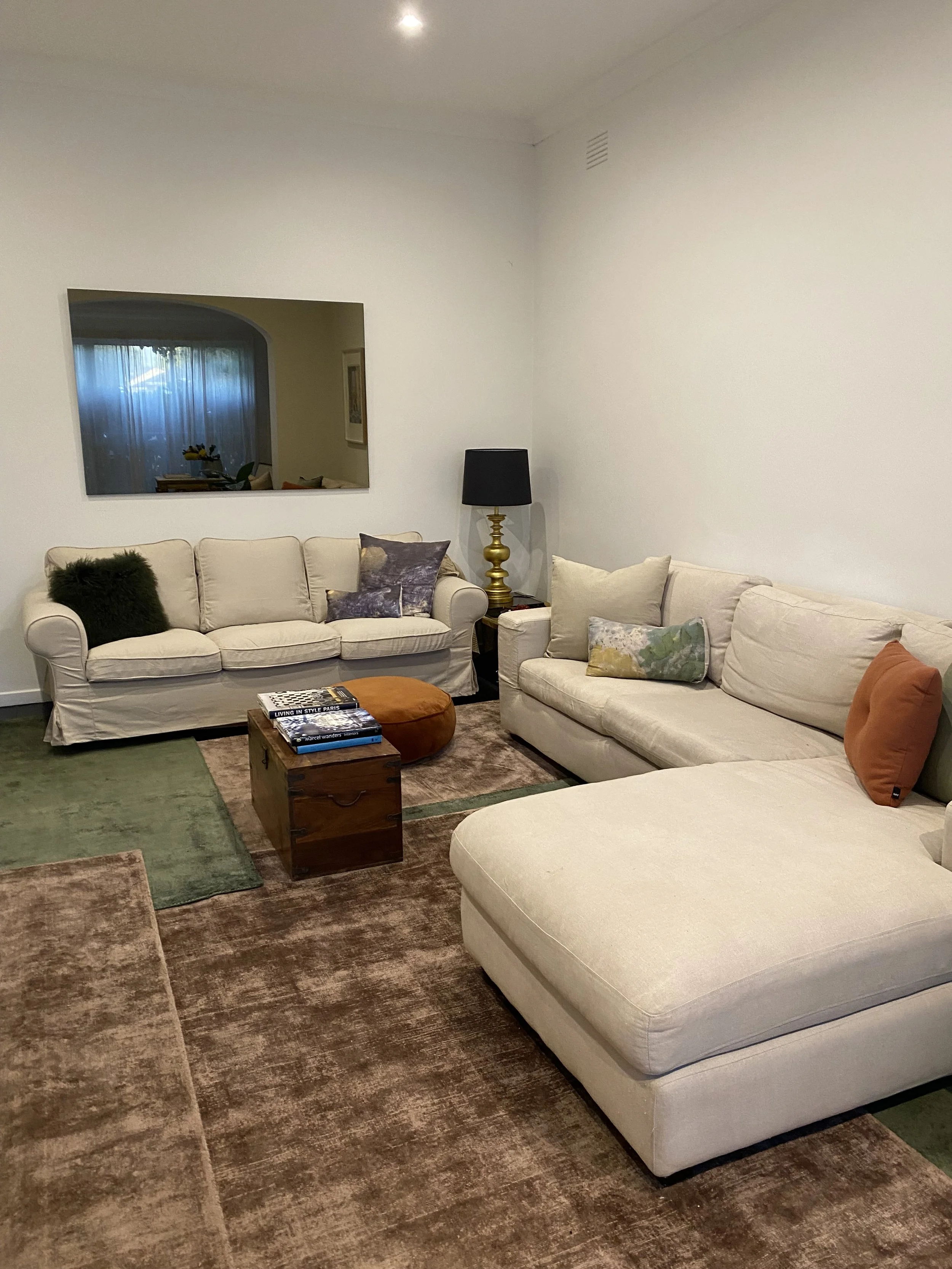 Living room with two beige sofas, a wooden coffee table with books, a black and gold table lamp, and colorful cushions. The room has a reflective wall mirror and a window with curtains.