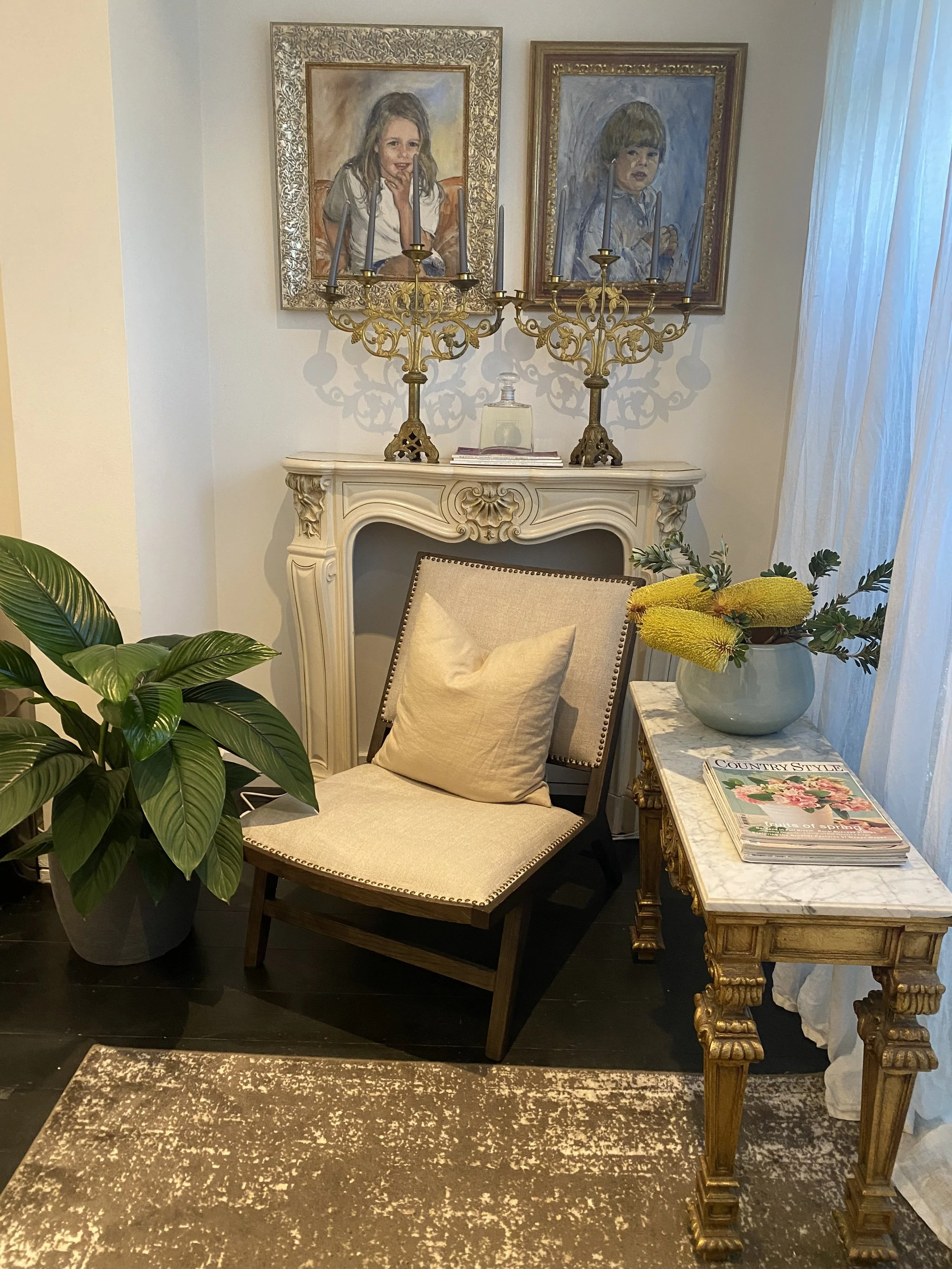 A cozy corner of a room featuring two framed paintings of children on the wall above a white ornate fireplace mantel. Two golden candle holders with candles sit on the mantel. To the left is a large green houseplant in a gray pot. In front of the fir
