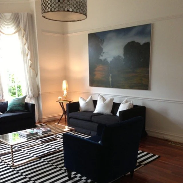 Living room with black sofas, white pillows, striped rug, glass coffee table, side table with lamp, large landscape painting, window with white curtains, and ceiling light fixture.