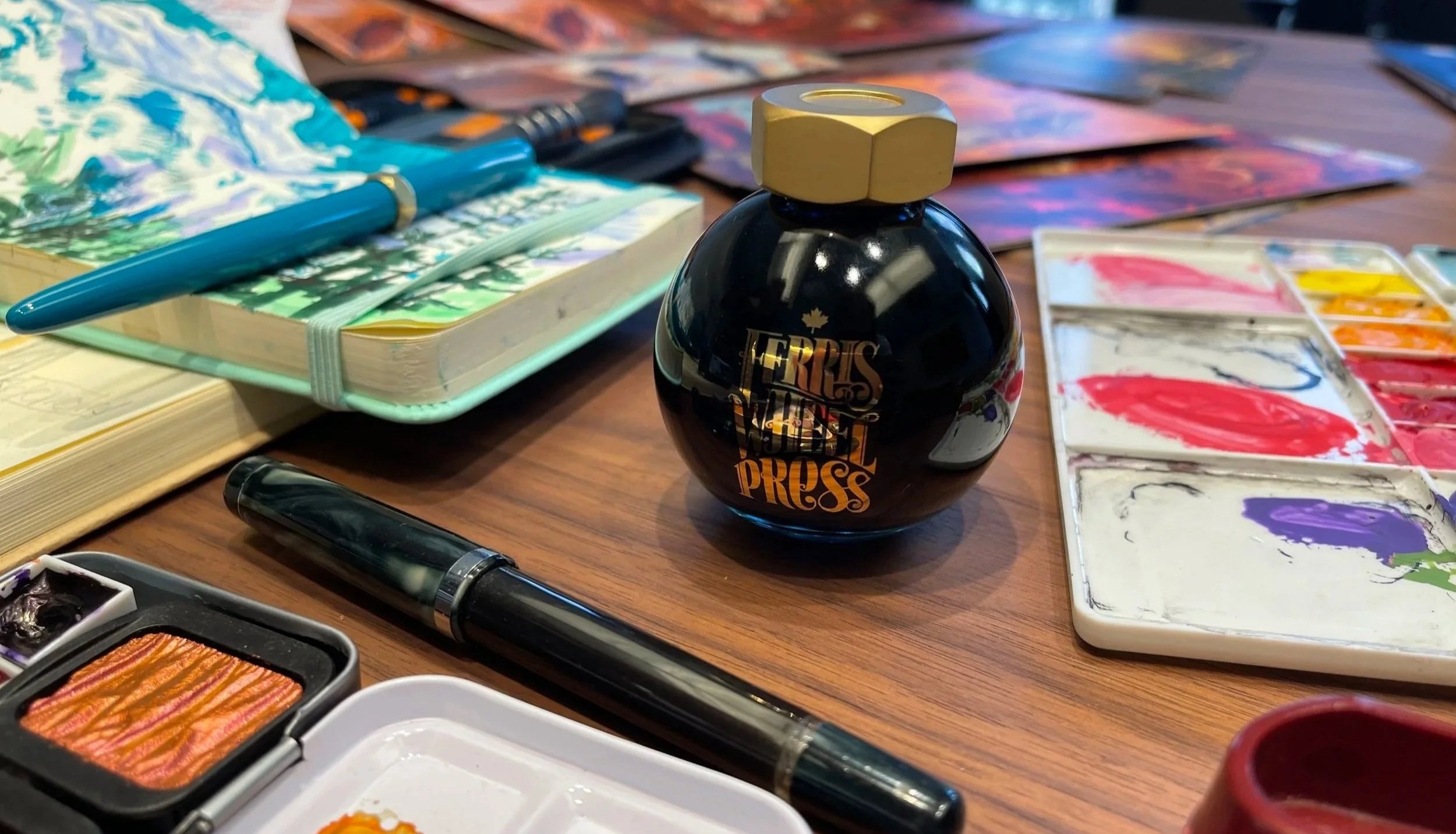 Art supplies on a wooden table, including a watercolor palette with paint, a black ink bottle labeled 'FERRIS WHEEL PRESS,' various brushes, pens, notebooks, and watercolor paper with painted swatches.
