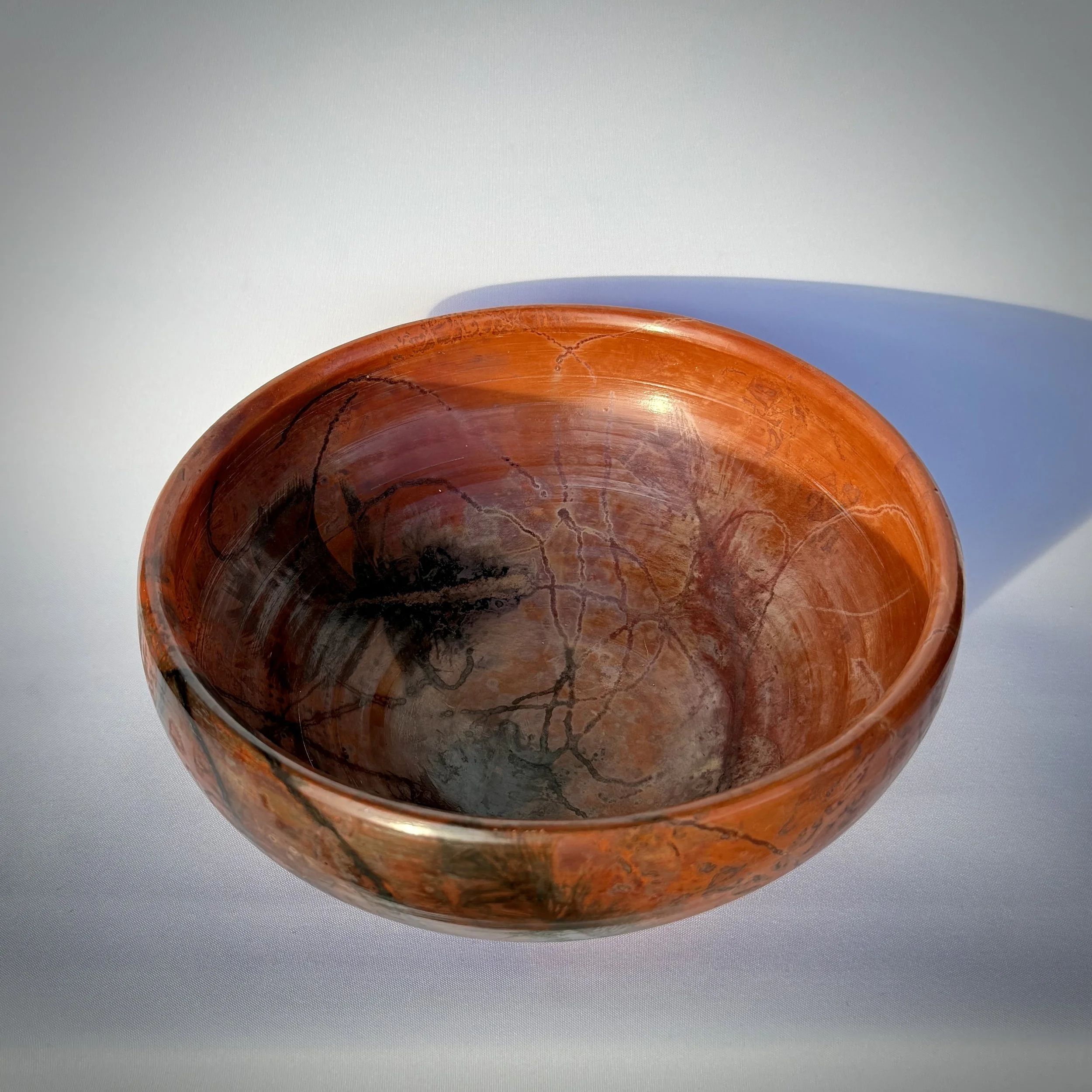 A polished, pit fired orange-brown ceramic bowl with visible vein-like patterns from wire and feathers inside, casting a shadow on a white surface.