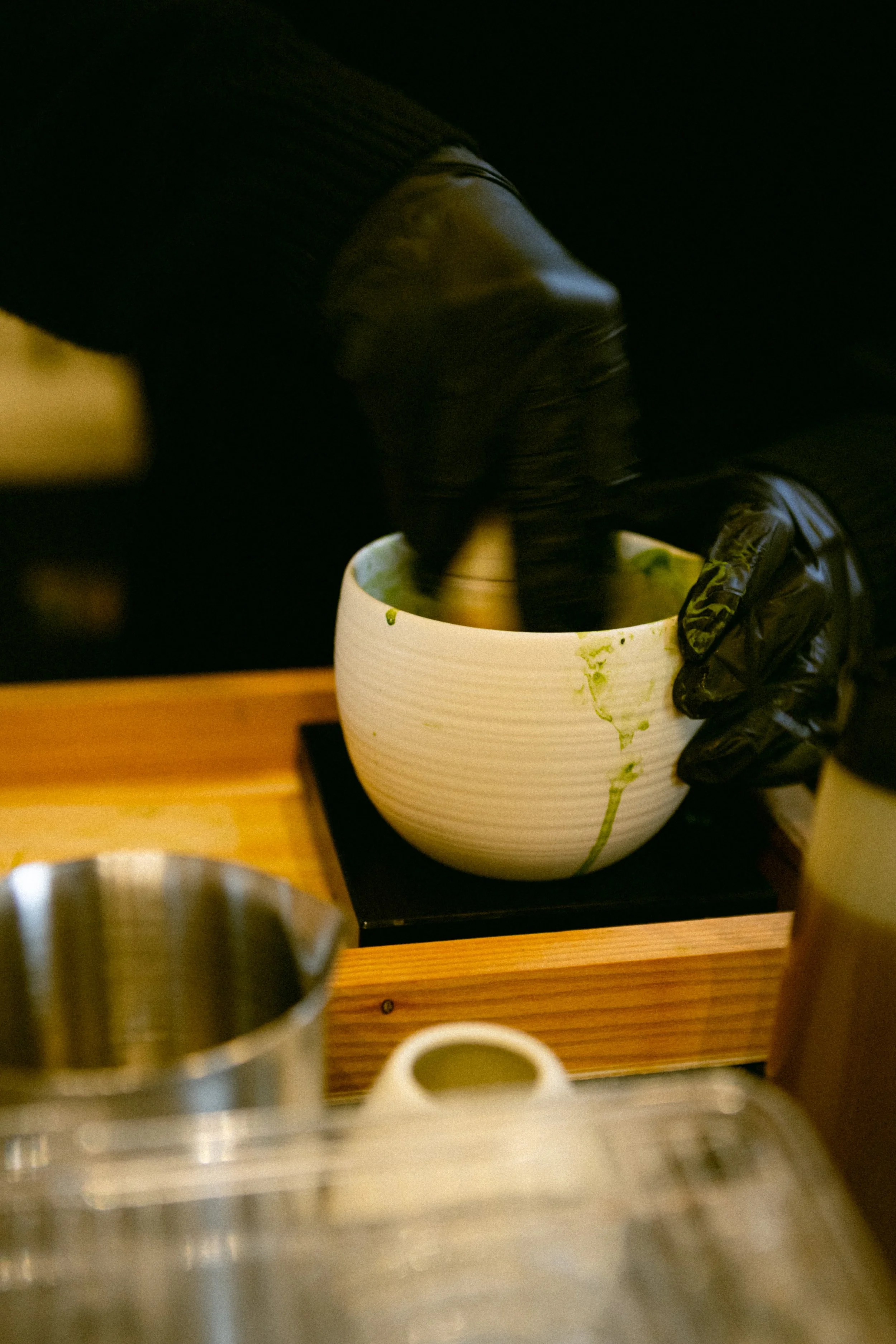 Person wearing black gloves mixing a green substance in a white bowl placed on a wooden stand.