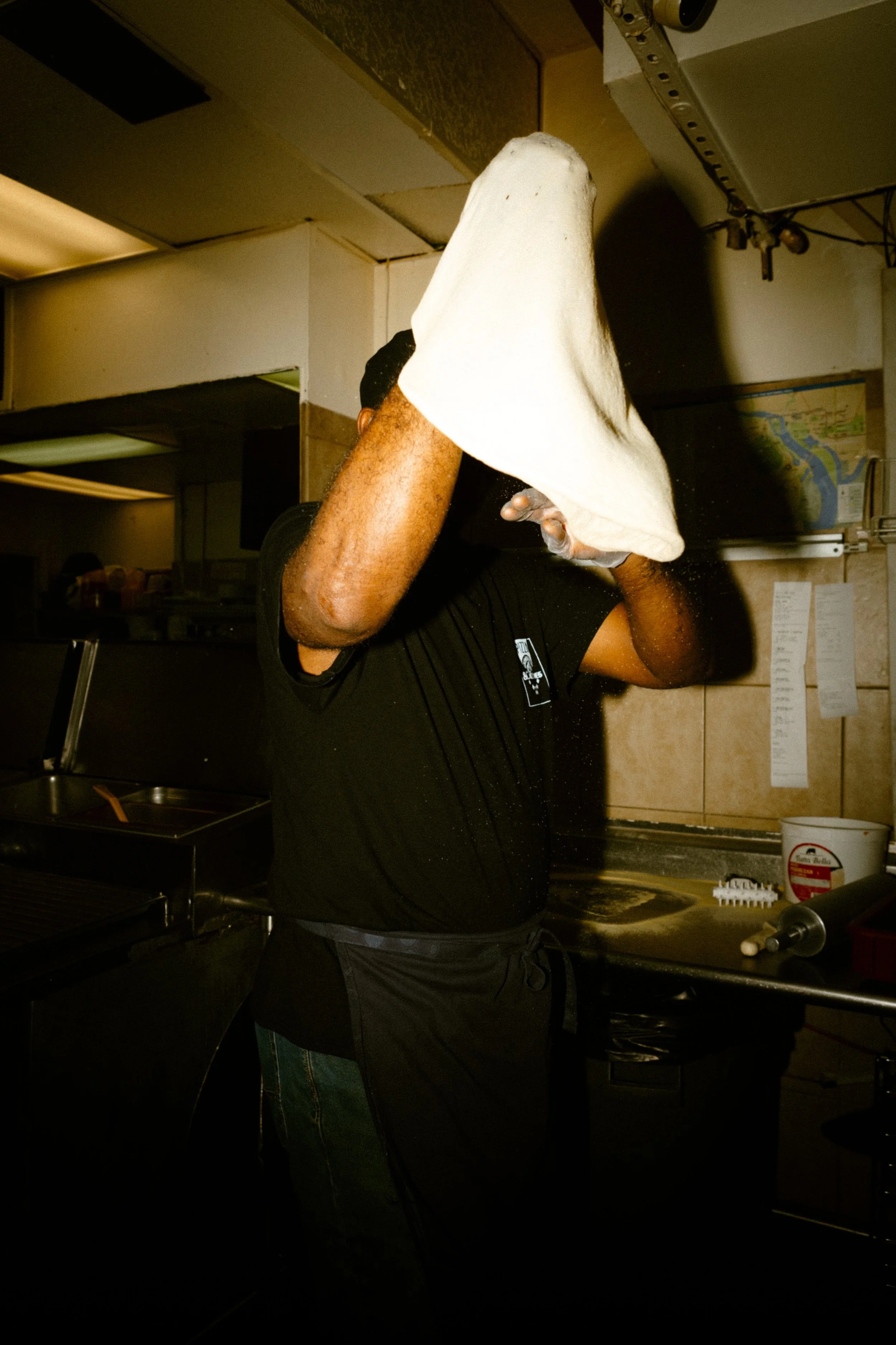 A person in a black T-shirt and apron, with raised arms, preparing or tossing pizza dough in a kitchen.
