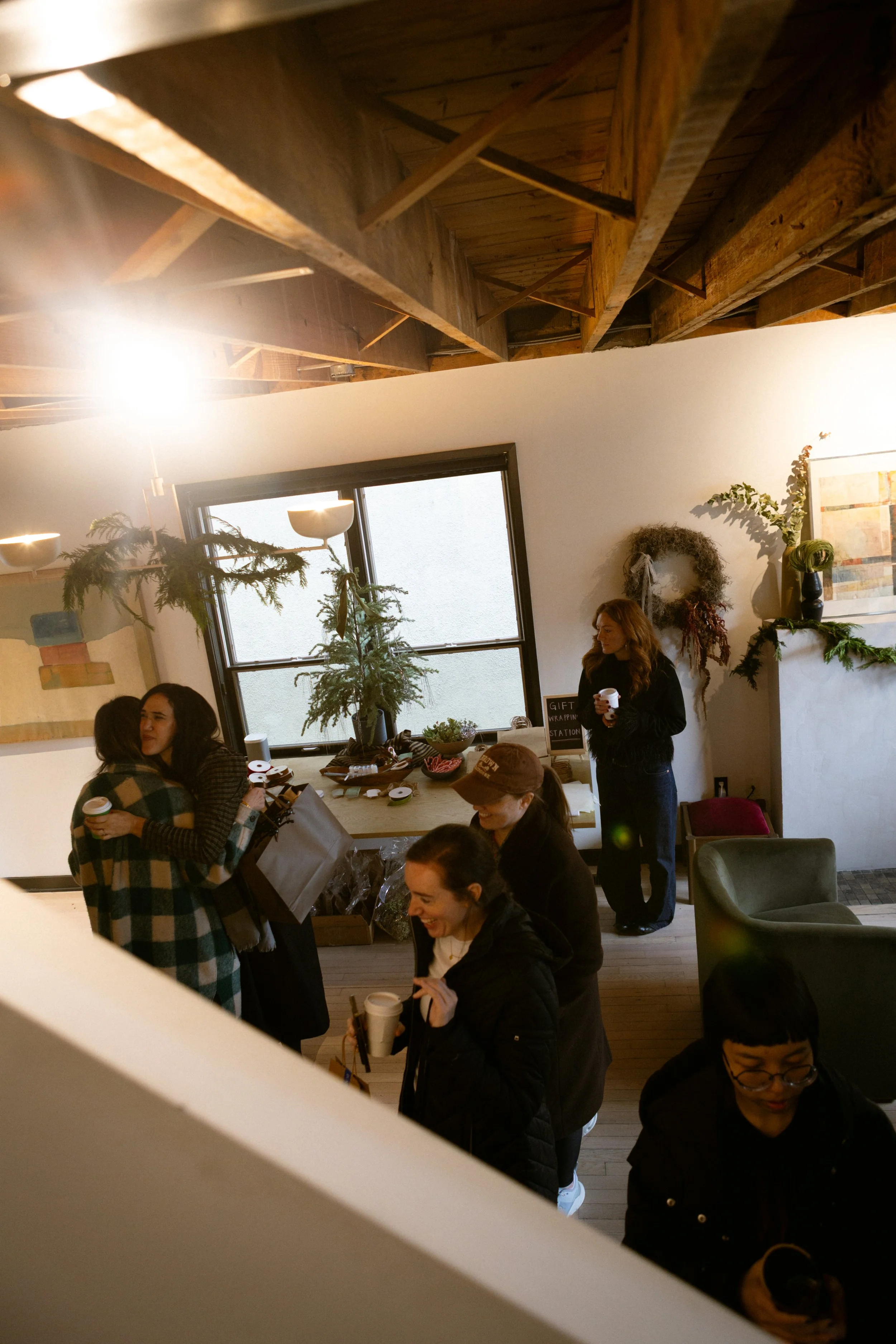People gathered in a cozy room, some hugging and holding cups, decorated with holiday greenery and artwork, during a festive event.