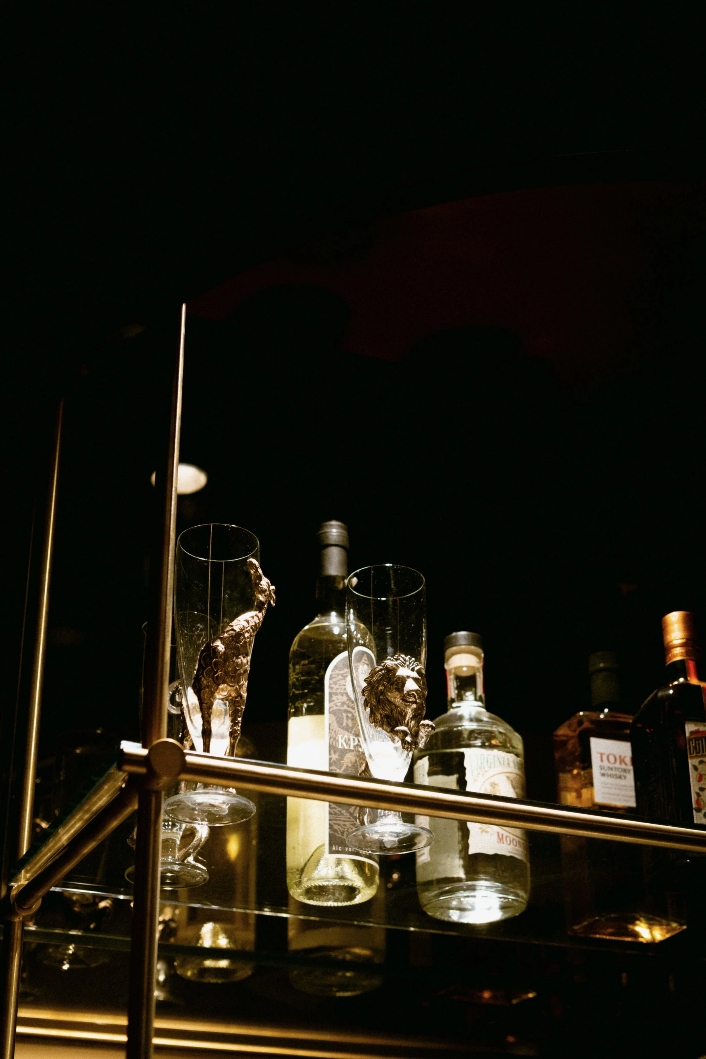 Display of liquor bottles and glassware with lion head details on glasses on a dark shelf.