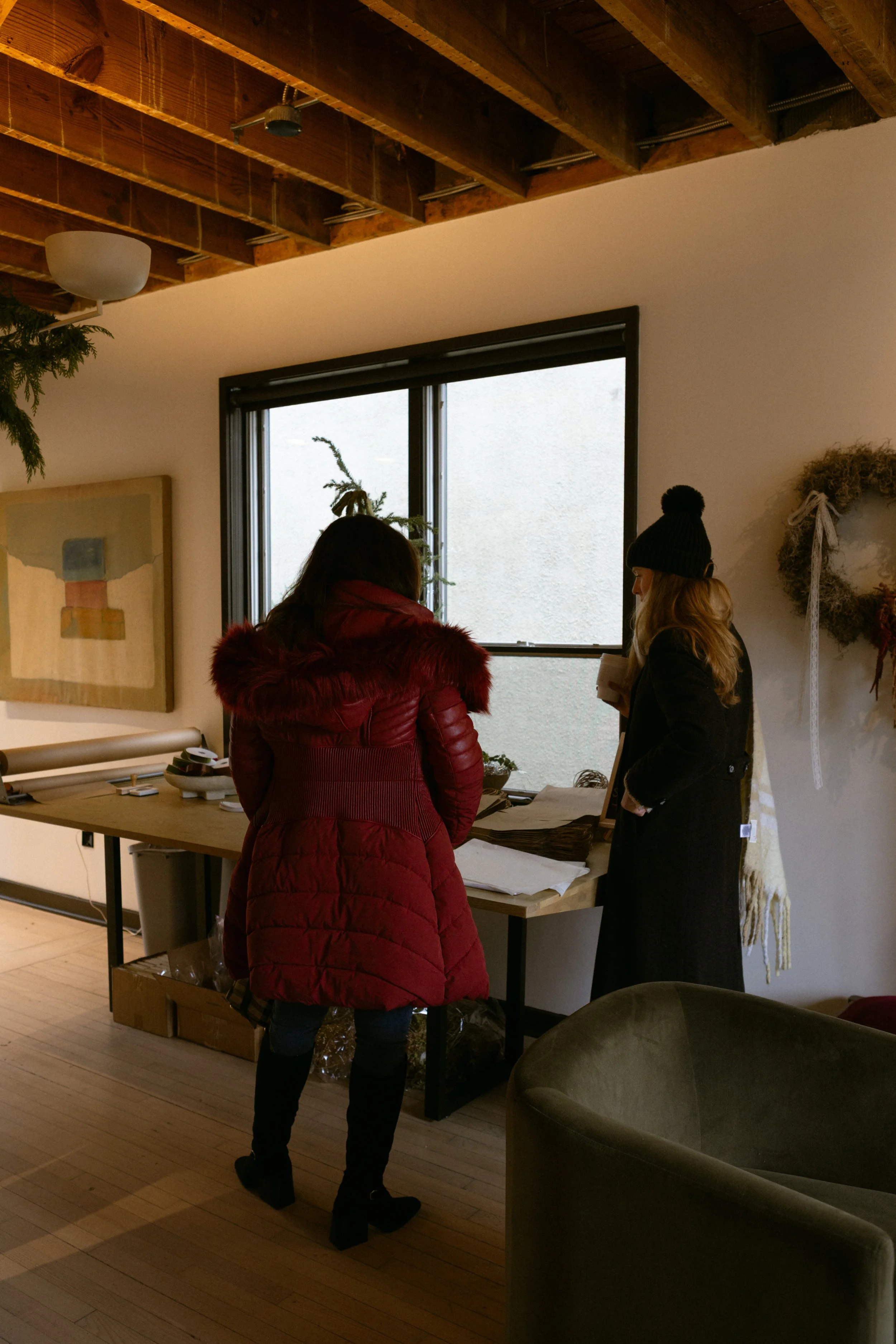 Two women in winter coats stand near a table inside a room with a wooden ceiling, a window, and holiday decorations.