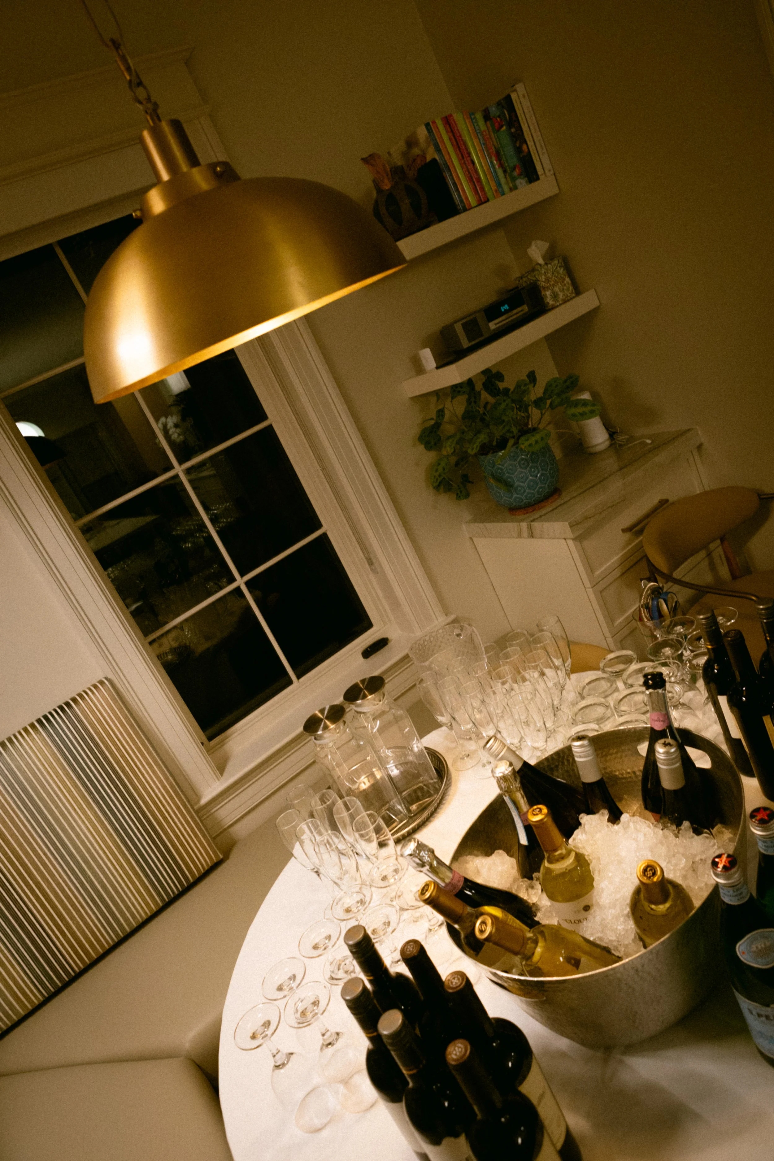 A round table with various bottles of beer, champagne glasses, and other glassware inside a warm-lit room, with a window and shelves in the background.