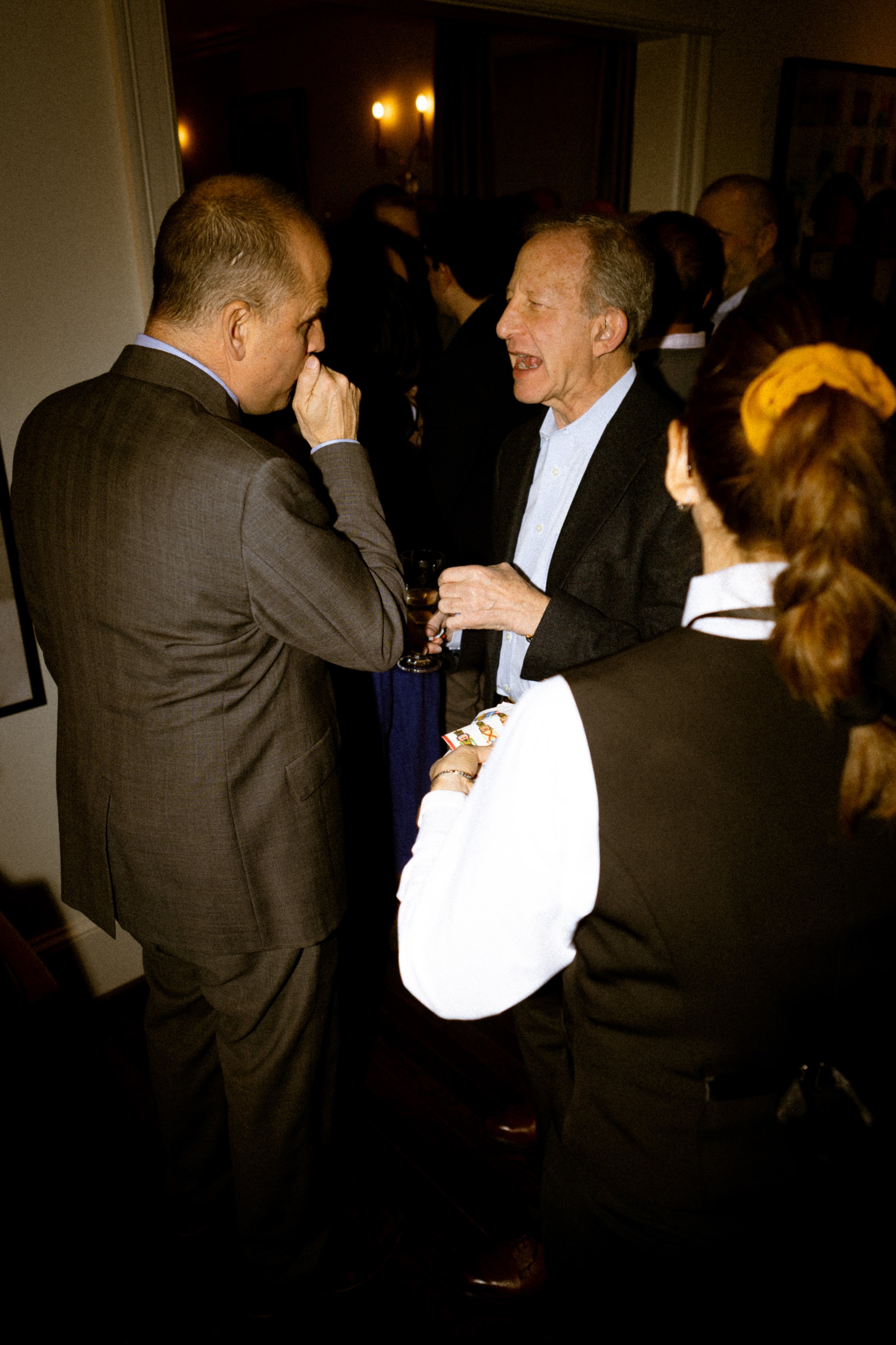 A group of people at a social gathering, with two men in conversation and a woman holding a glass, in an indoor setting with warm lighting.