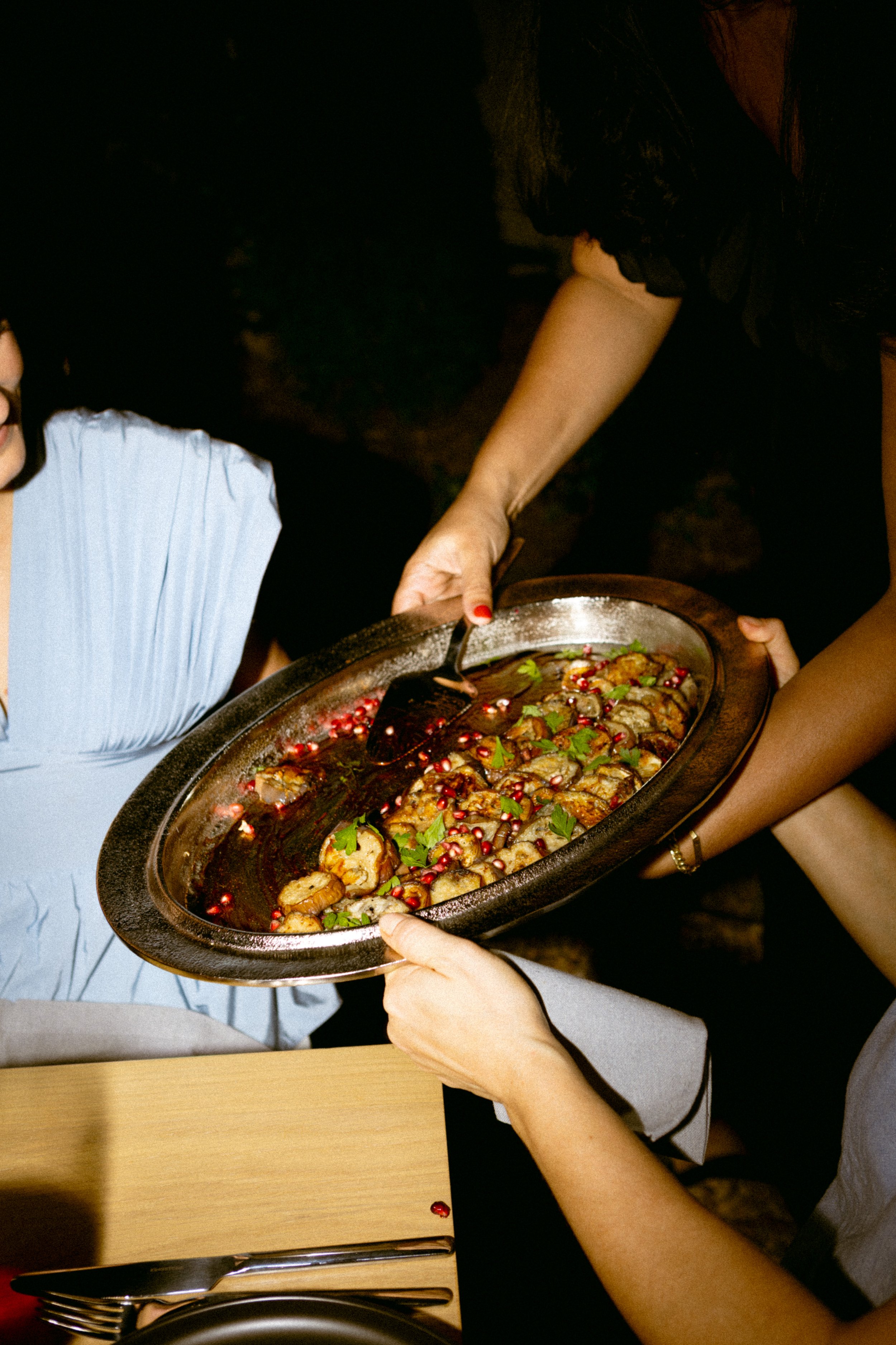 People serving and receiving a dish of food garnished with pomegranate seeds and herbs at a dinner.
