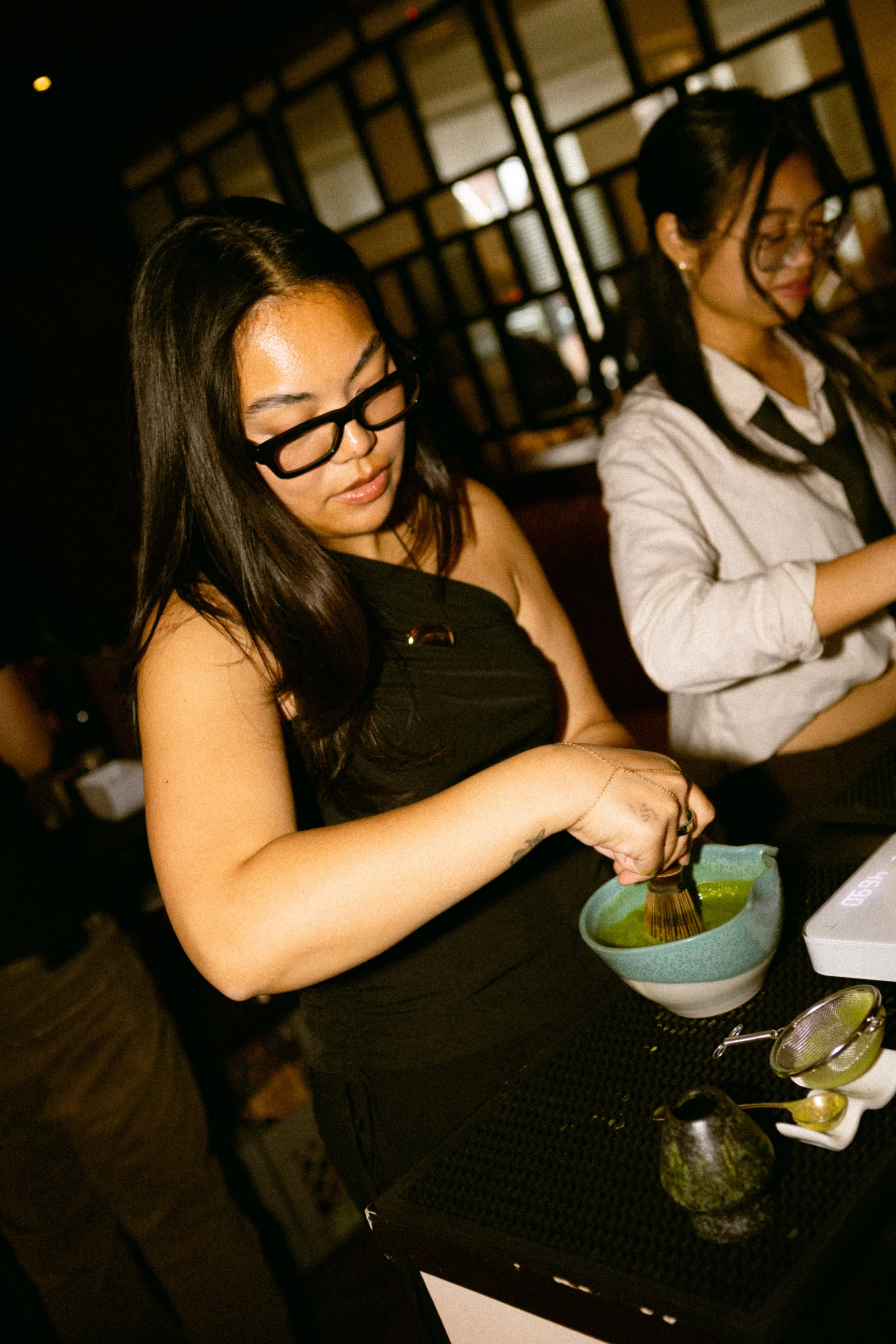 A woman with long dark hair, black glasses, and a black sleeveless top is whisking matcha tea in a ceramic bowl. Another woman with long dark hair, wearing glasses, a white shirt, and black tie, is sitting next to her at a table in a tea or coffee sh
