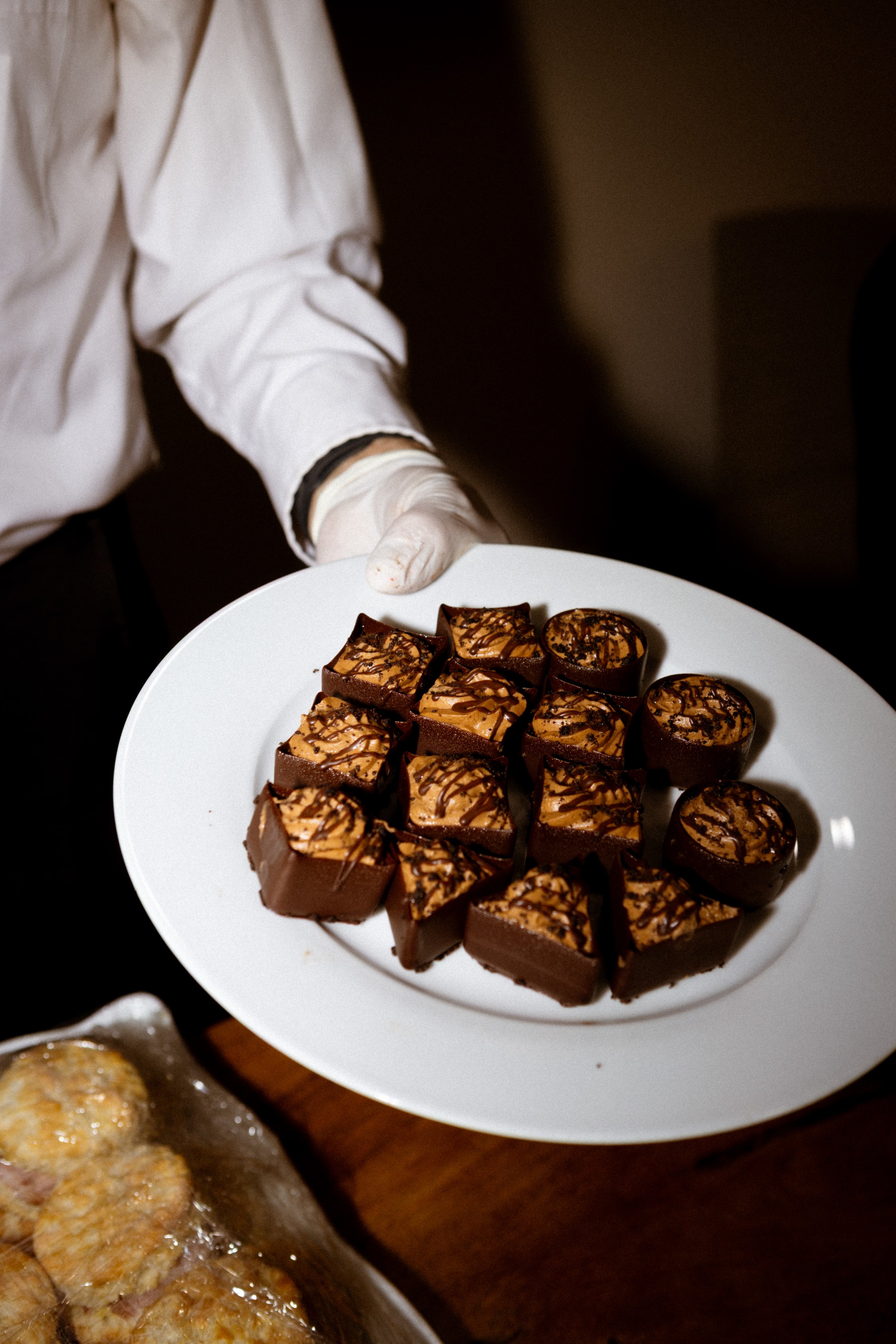 Person in white shirt and gloves holding white plate with assorted chocolate desserts topped with caramel and drizzled with chocolate.