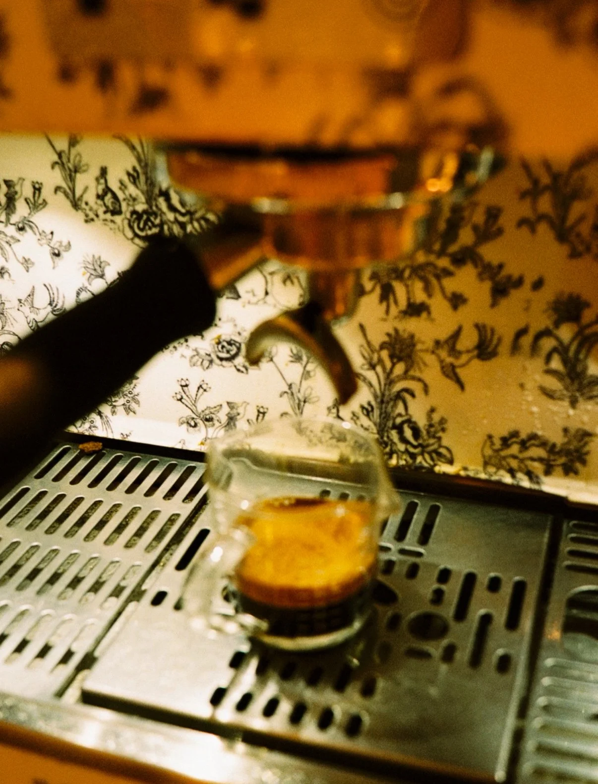 A person is pouring a shot of espresso into a small glass cup on a metal drip tray, with a decorative floral wallpaper in the background.